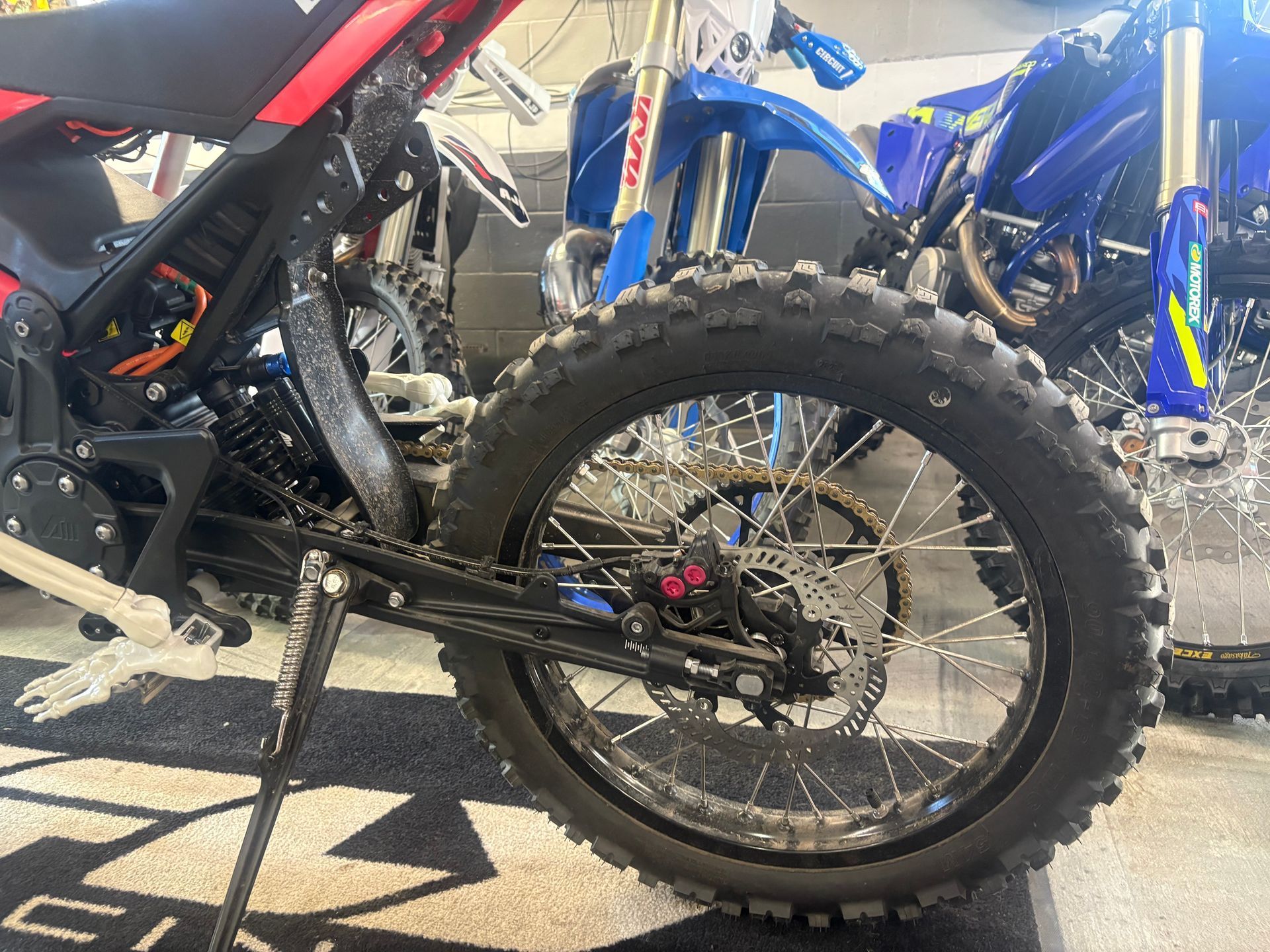 Dirt bike rear wheel and frame with black and red bodywork, parked on pavement beside blue bikes.
