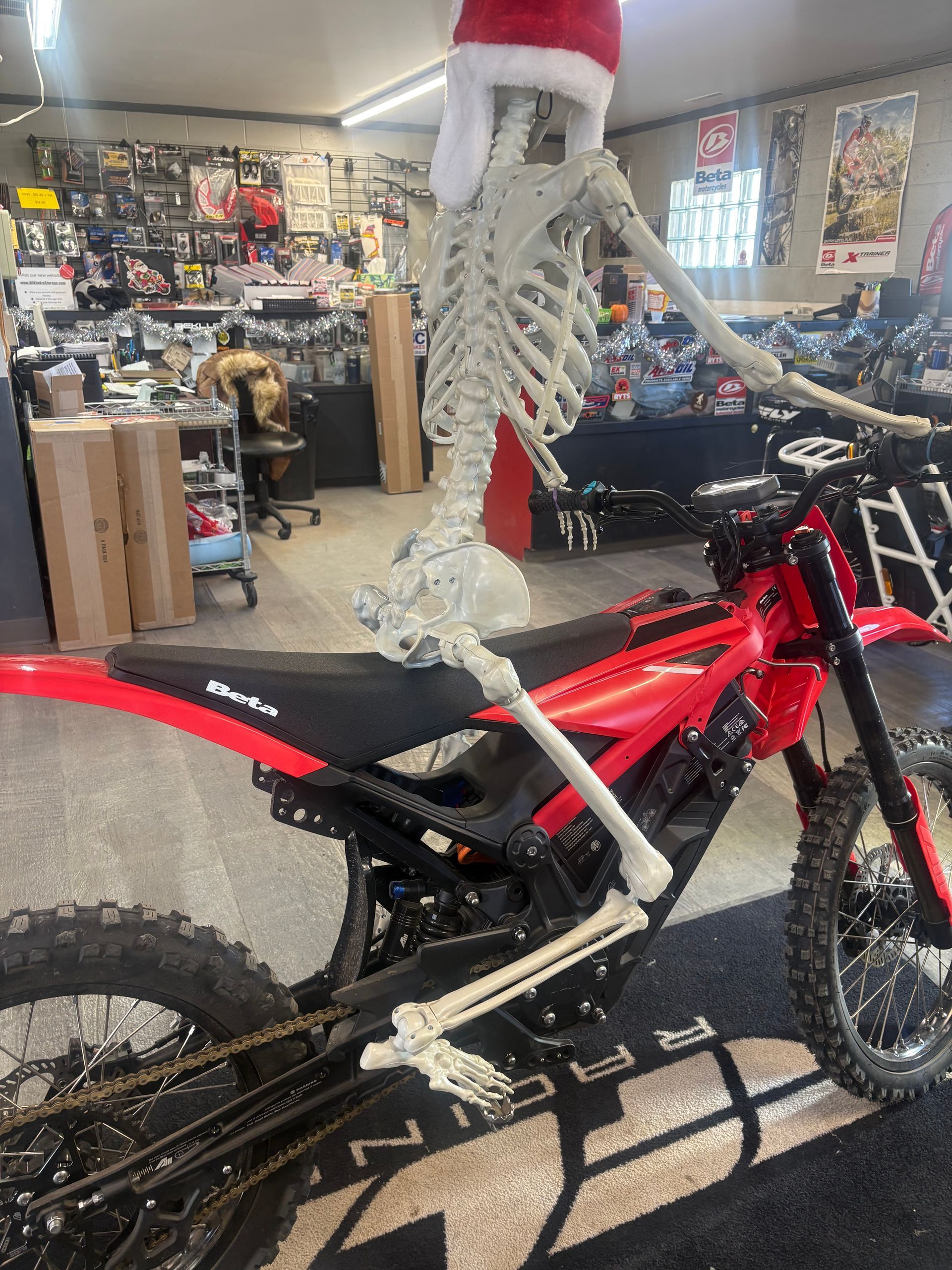 Skeleton on a red dirt bike inside a shop, with posters and motorcycle gear in the background