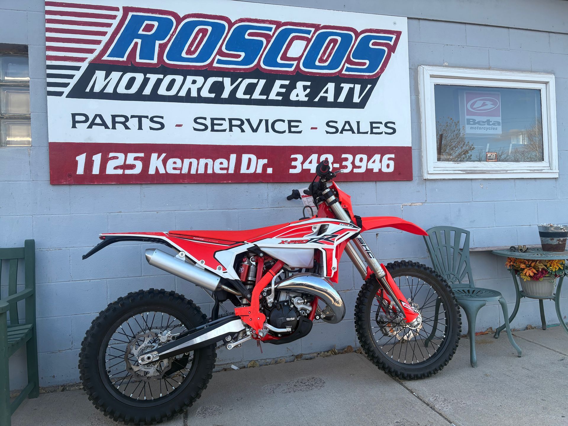 Red dirt bike parked outside Rosco's Motorcycle & ATV shop sign in front of a gray building