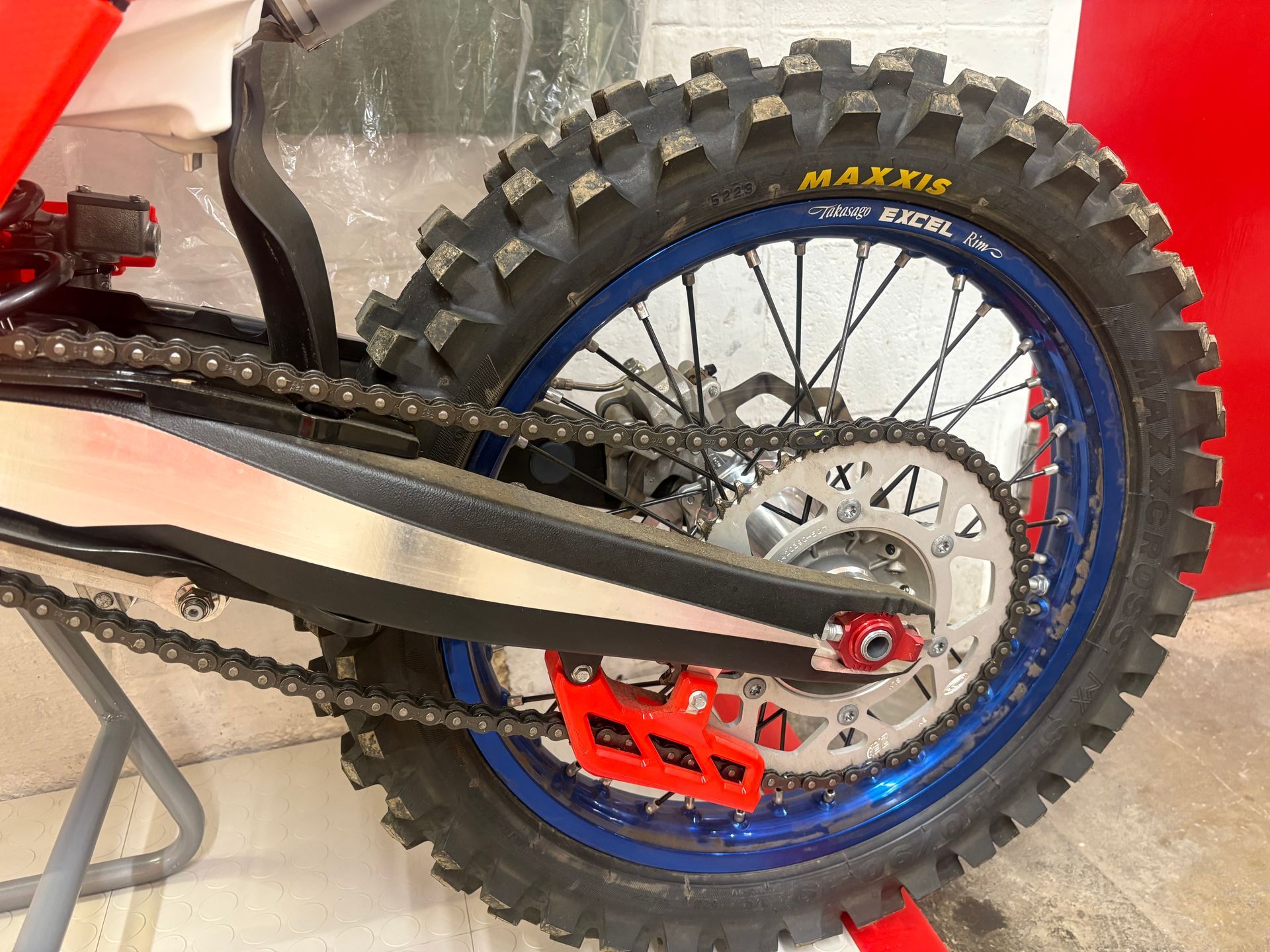Close-up of a dirt bike’s rear wheel, chain, and red footpeg with a blue rim and knobby tire.