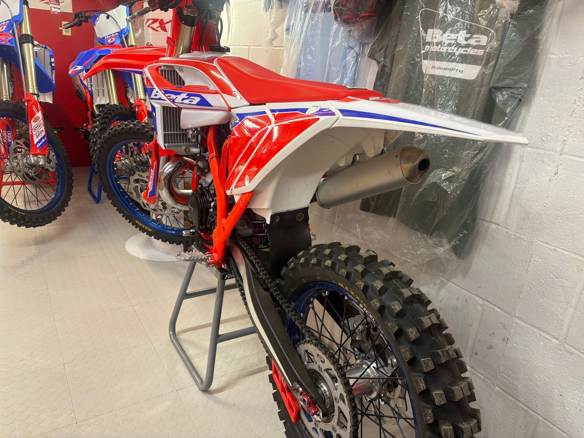 Red and blue dirt bike in a workshop, shown from the rear with knobby tire and raised stand