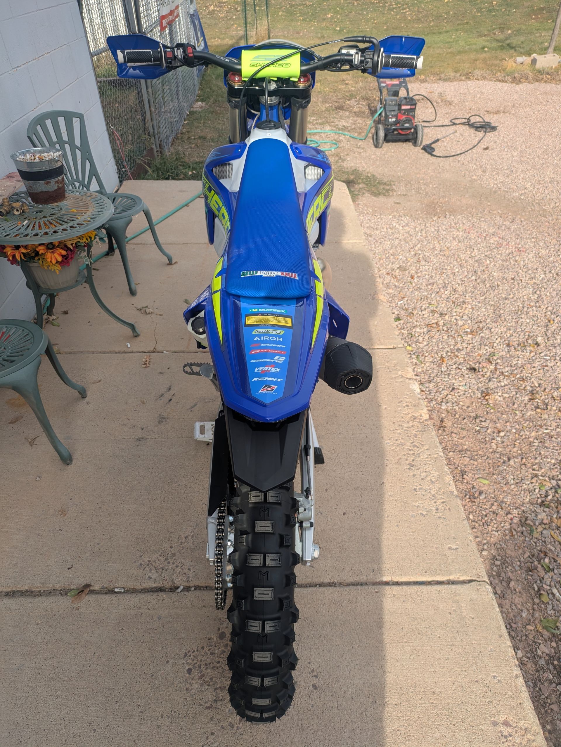 Blue dirt bike parked on a concrete path beside a gravel yard, viewed from the front.