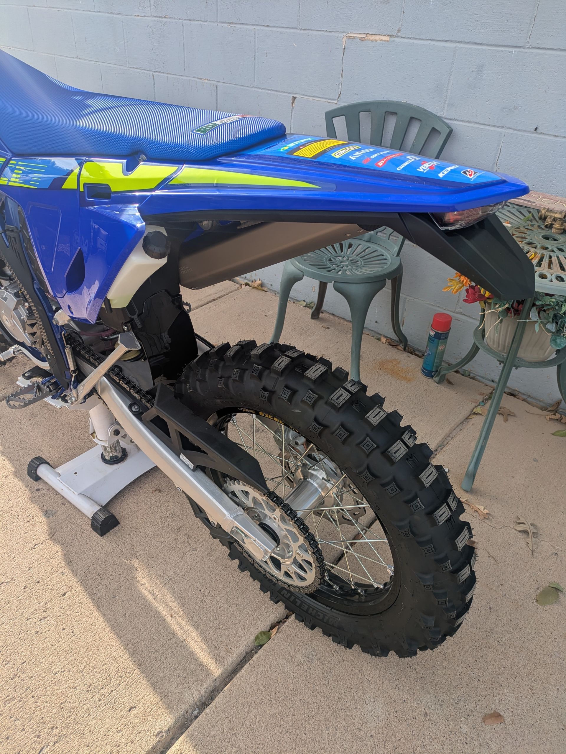 Blue dirt bike rear wheel and seat in a patio area, with a chair and bottle in the background