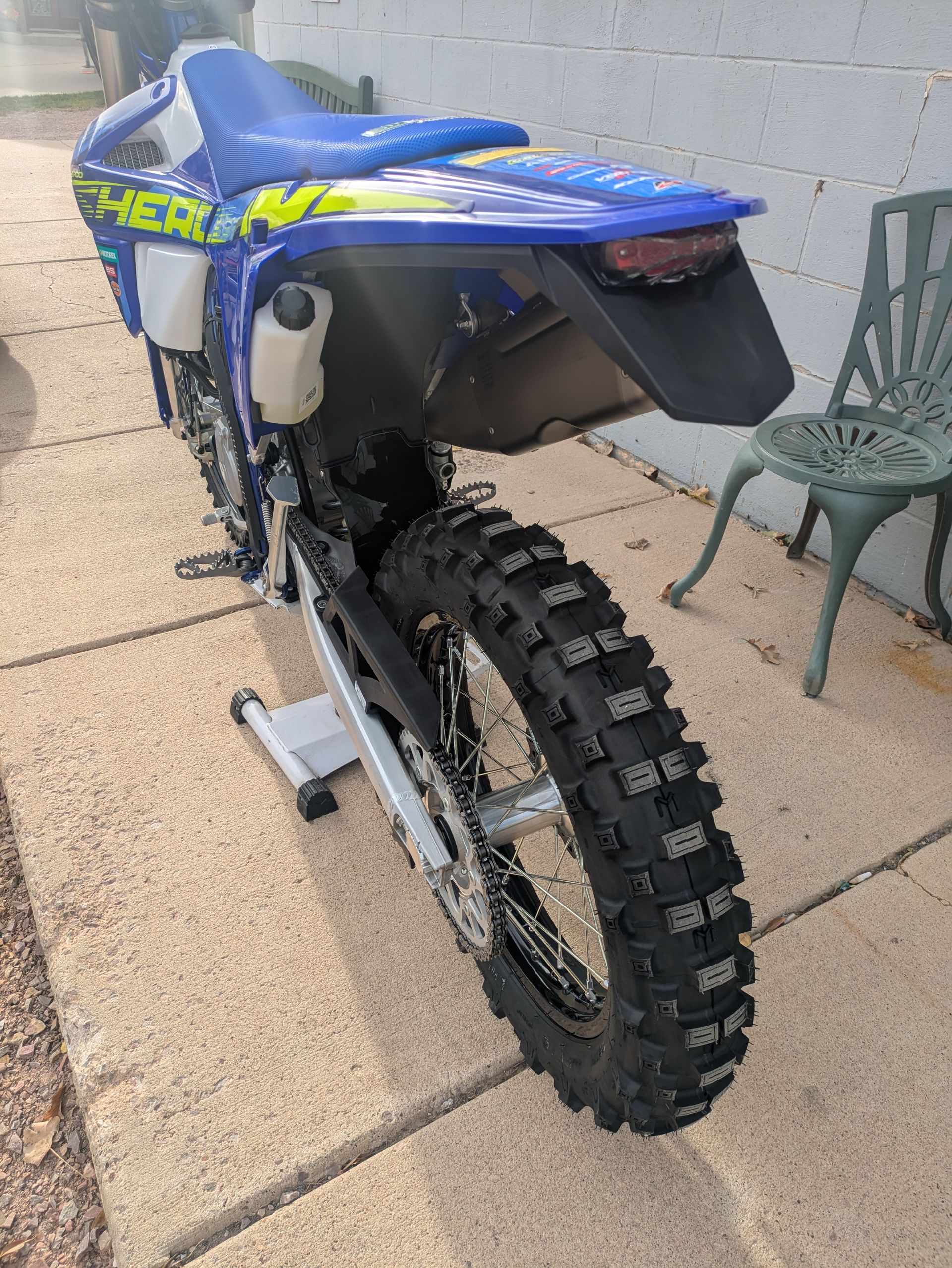 Blue and green dirt bike parked on a patio beside a chair, rear wheel in view