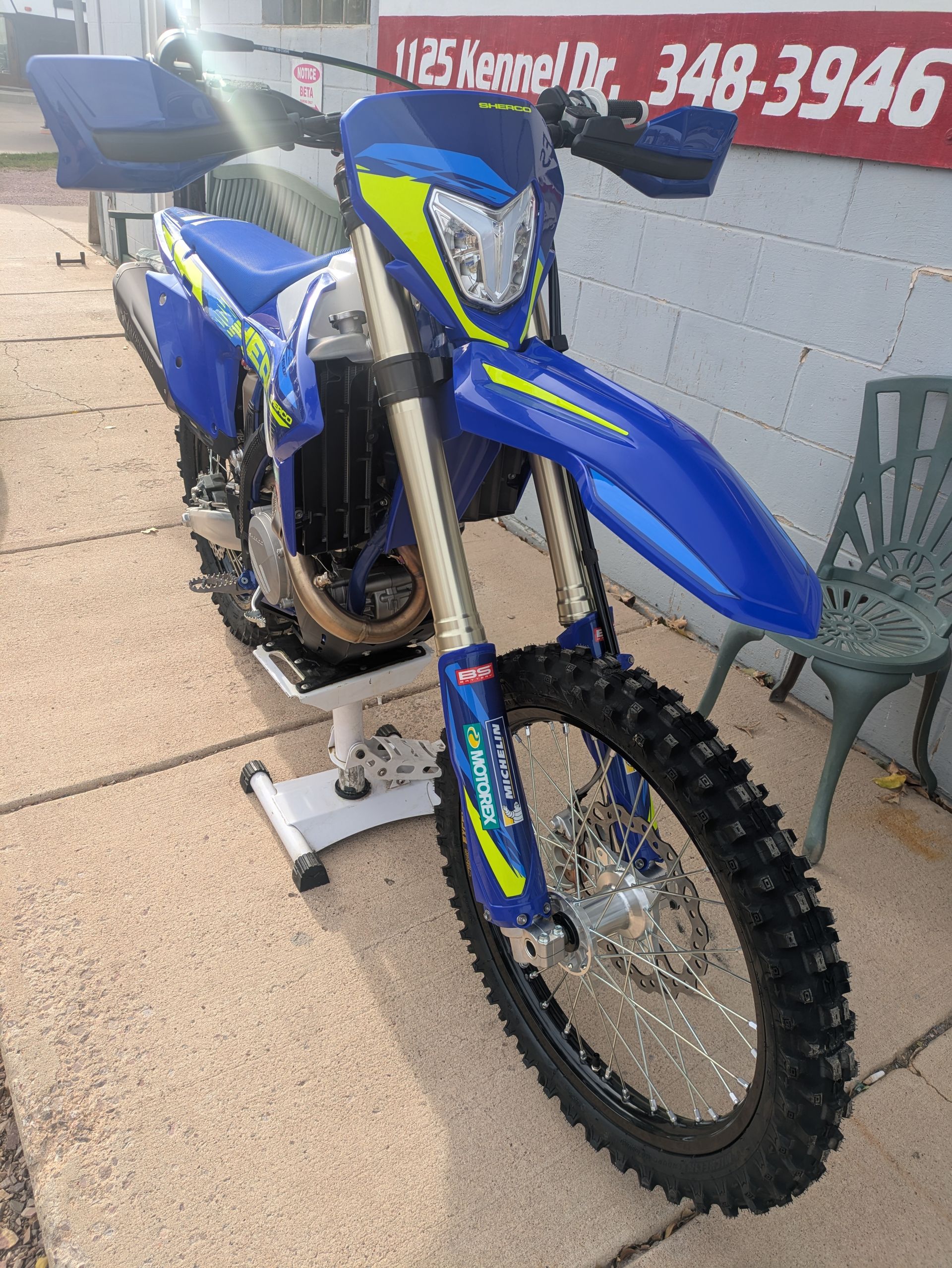 Blue dirt bike on a stand outdoors beside a wall and chair