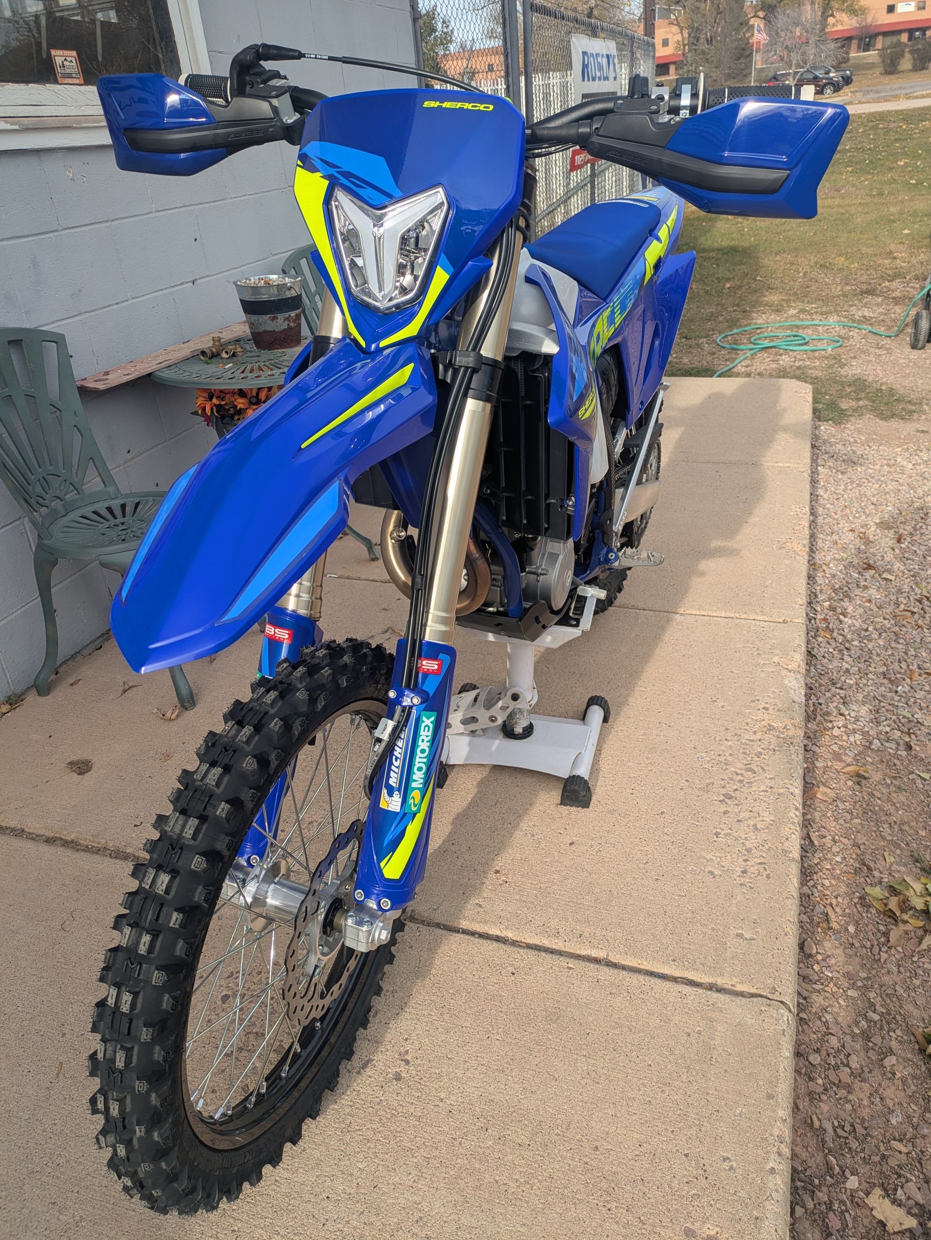 Blue dirt bike parked on a sidewalk, front view with neon green accents and knobby tires