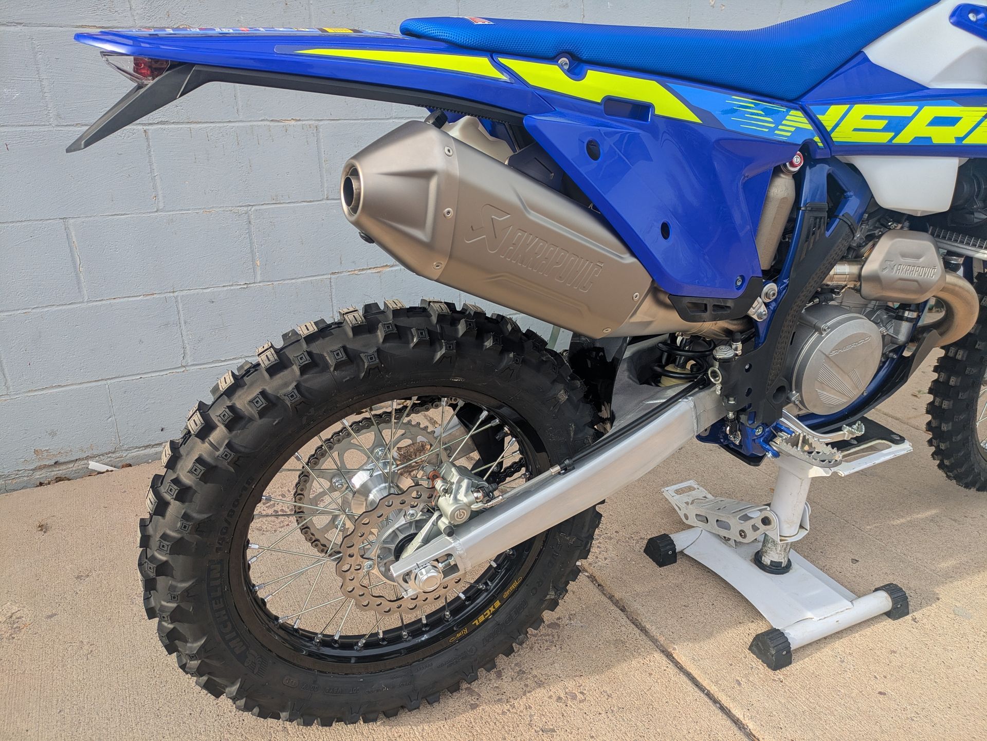 Blue dirt bike on a stand, rear wheel and exhaust visible, parked beside a gray wall.