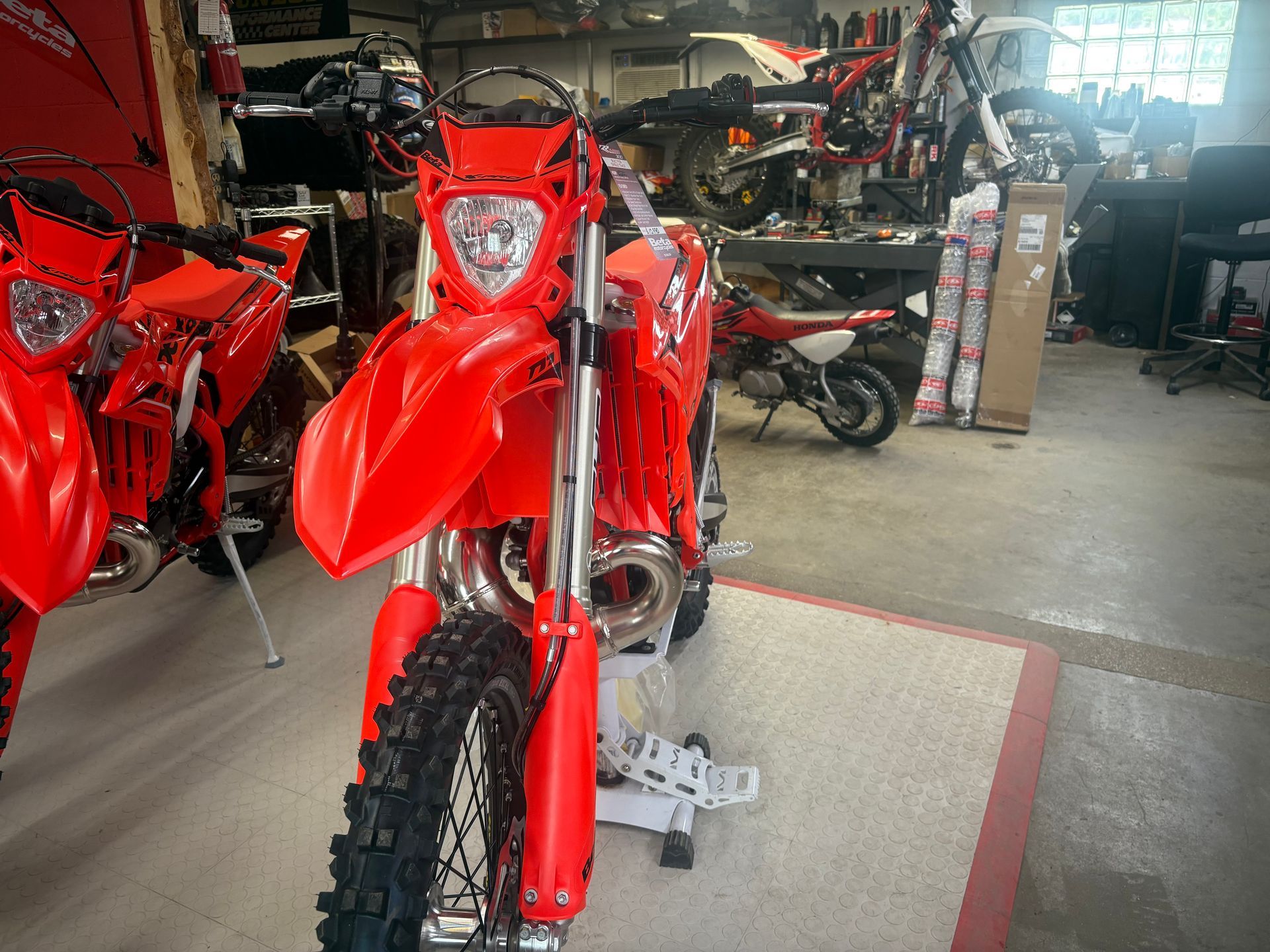 Red dirt bike in a motorcycle workshop, with other bikes and tools in the background