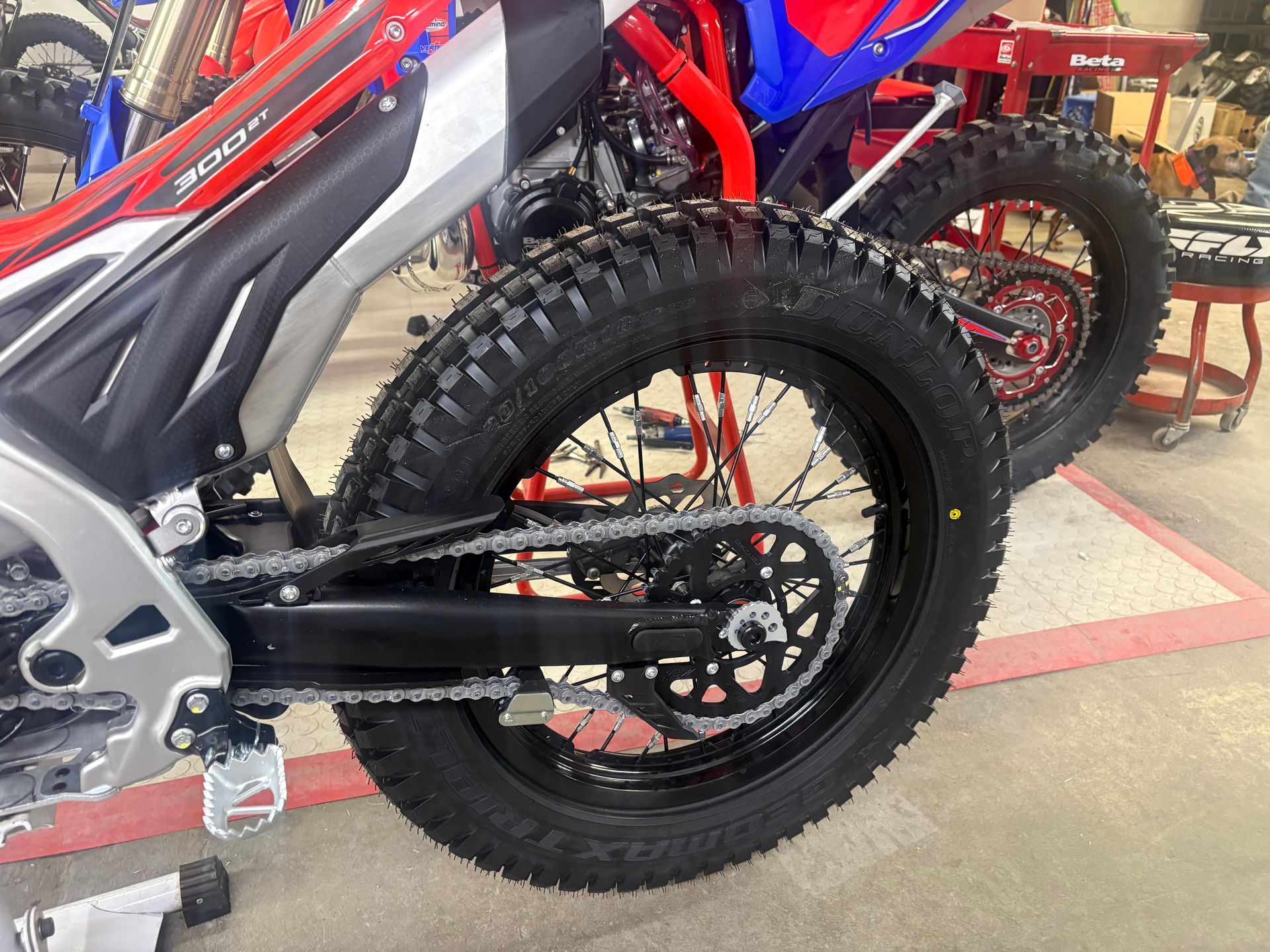 Close-up of a dirt bike's rear wheel, chain, and swingarm in a garage, with another bike in the background