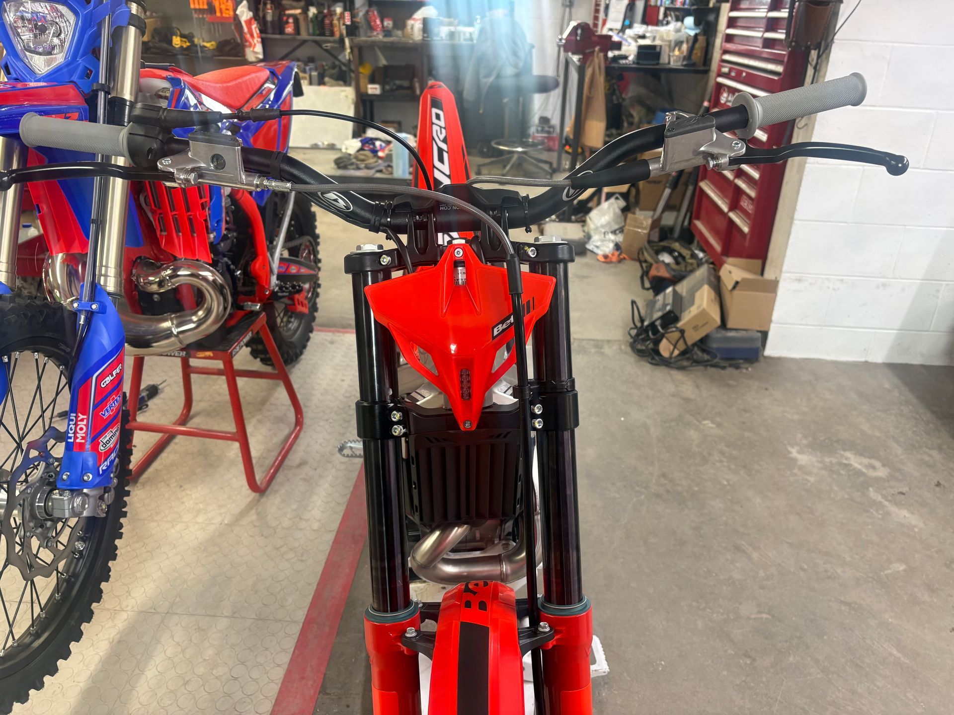 Red dirt bike in a workshop, with blue bikes on both sides and a concrete floor.