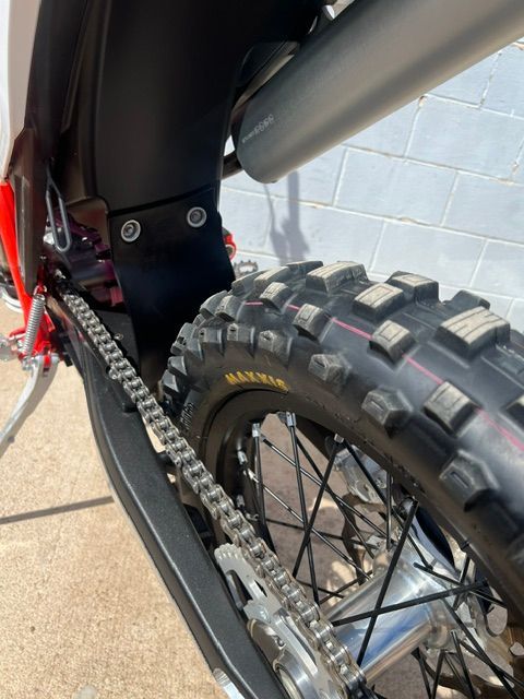 Close-up of a dirt bike rear wheel, chain, and suspension with a knobby tire against a gray wall