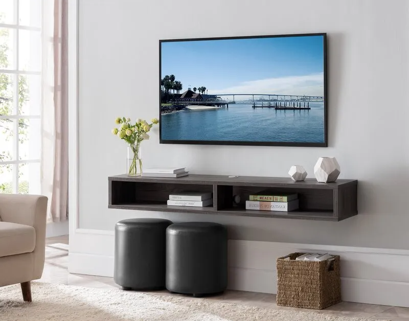 A TV mounted above a floating gray shelf with decor, two black ottomans, and a woven basket in a living room.