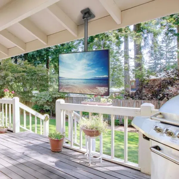 An outdoor TV mounted on a patio ceiling, showing a beach scene. Deck with a grill, railing, and greenery.