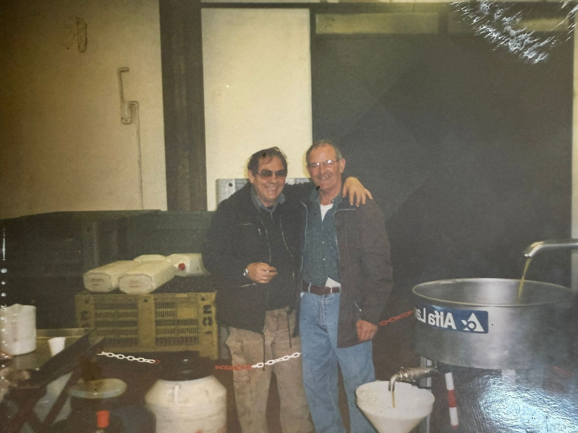 Due persone in piedi insieme in una stanza poco illuminata, accanto a barili e secchi, sorridono alla macchina fotografica.