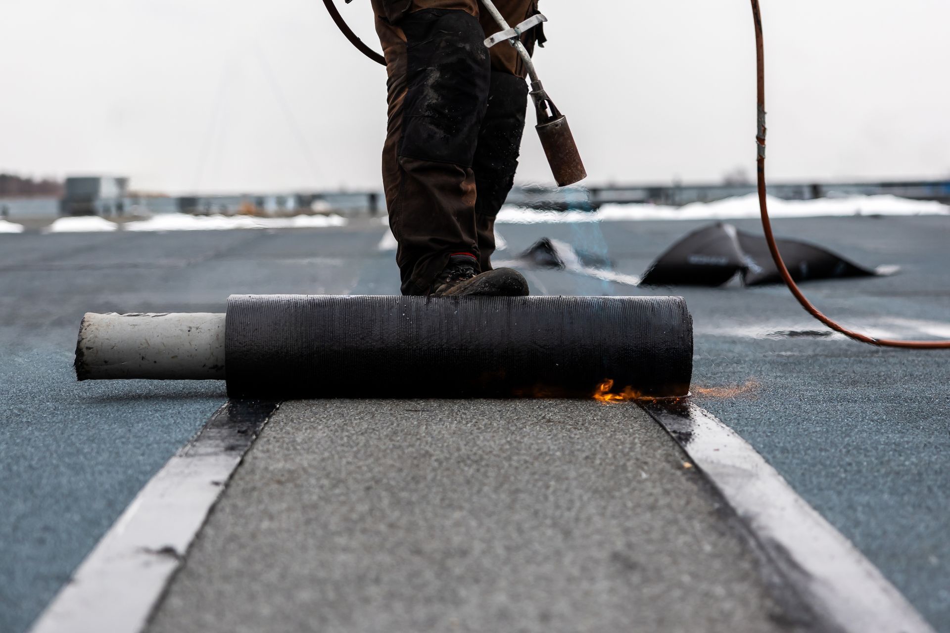 Worker torching roofing material on a flat roof, rolling out a black waterproof membrane.