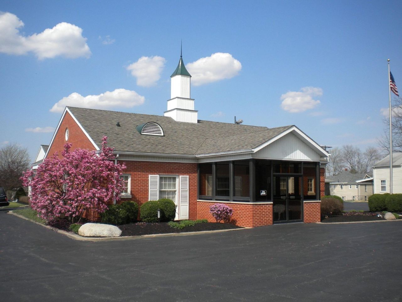 Small brick civic building with white steeple, pink flowering tree, and American flag on a sunny day