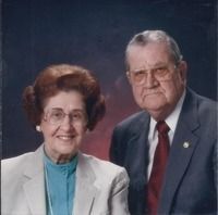 Two people posed together against a dark studio backdrop, wearing formal attire and smiling slightly.
