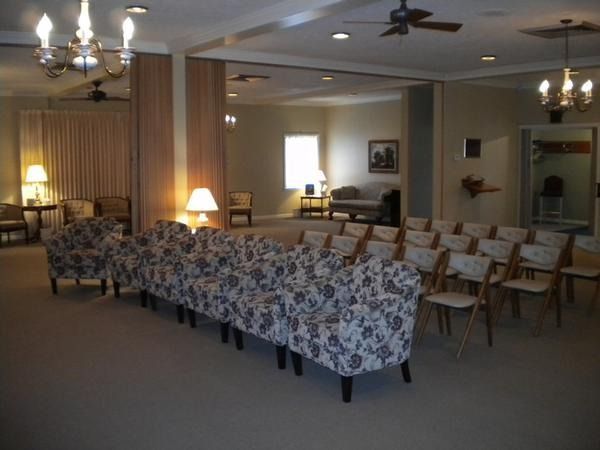 Empty community room with patterned sofas, rows of chairs, lamps, and ceiling fans under warm lighting