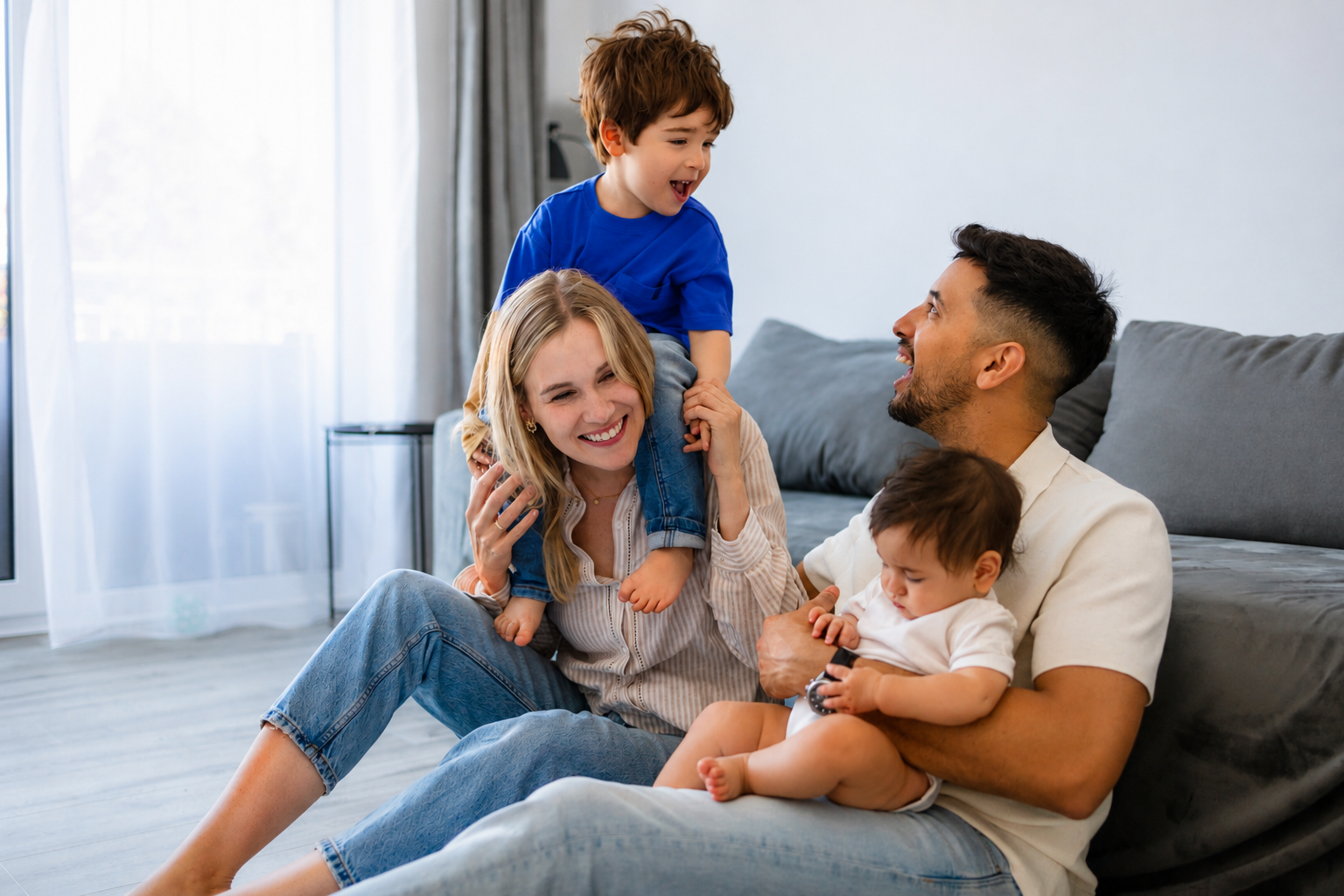 Family sitting on a couch, smiling and playing with two young children in a bright living room