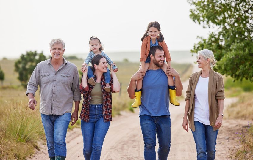 Family walking together on a dirt path, with two children riding on adults’ shoulders in a sunny rural field