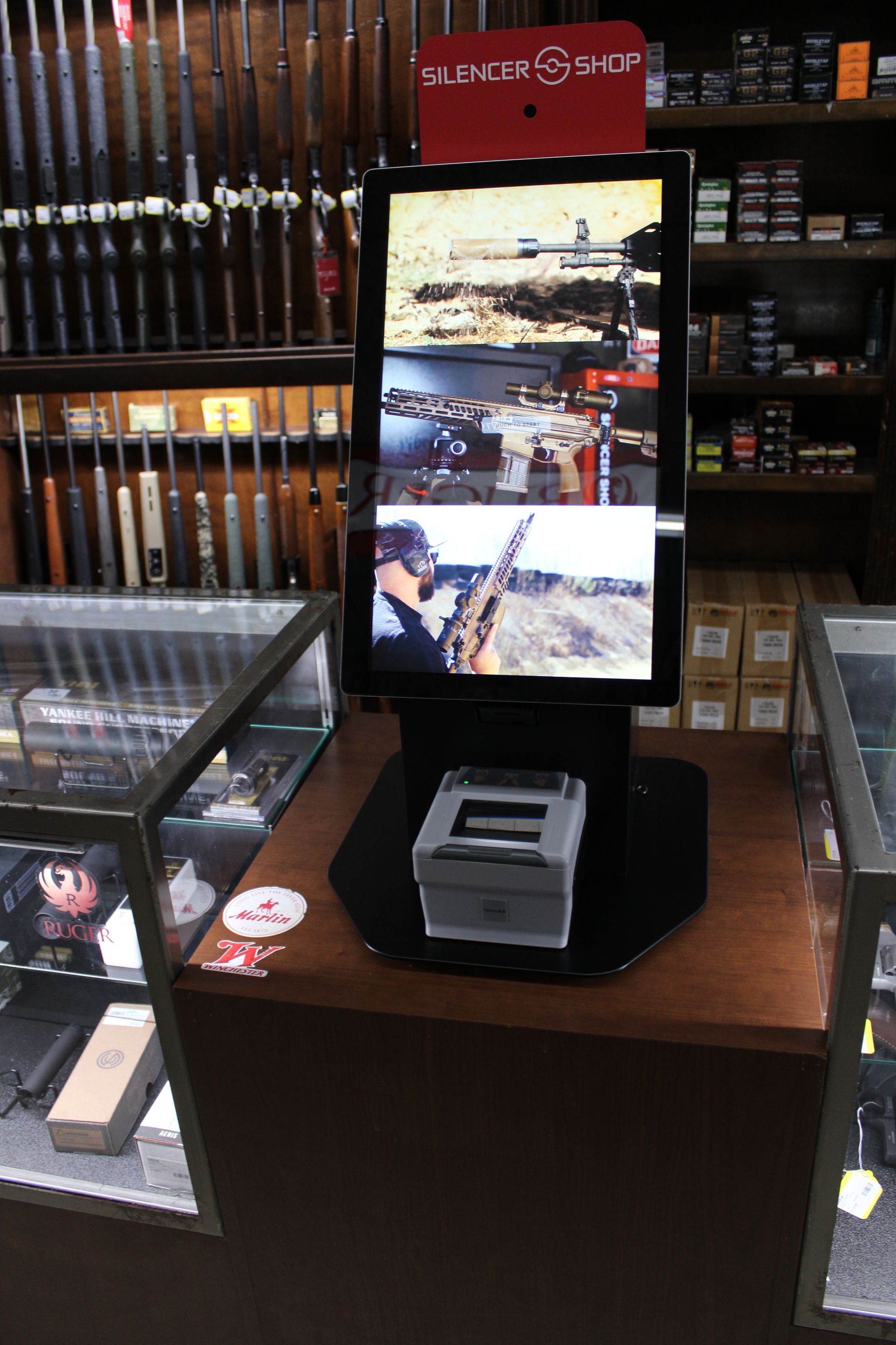 Vertical digital display in a retail store, showing a hunting scene amid display cases and gun racks
