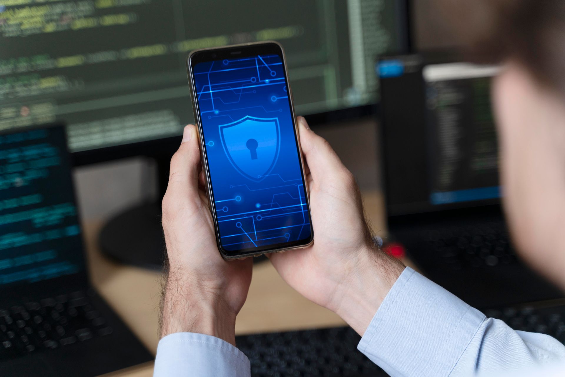 Mãos segurando um smartphone com uma película de segurança azul, em frente a telas de computador