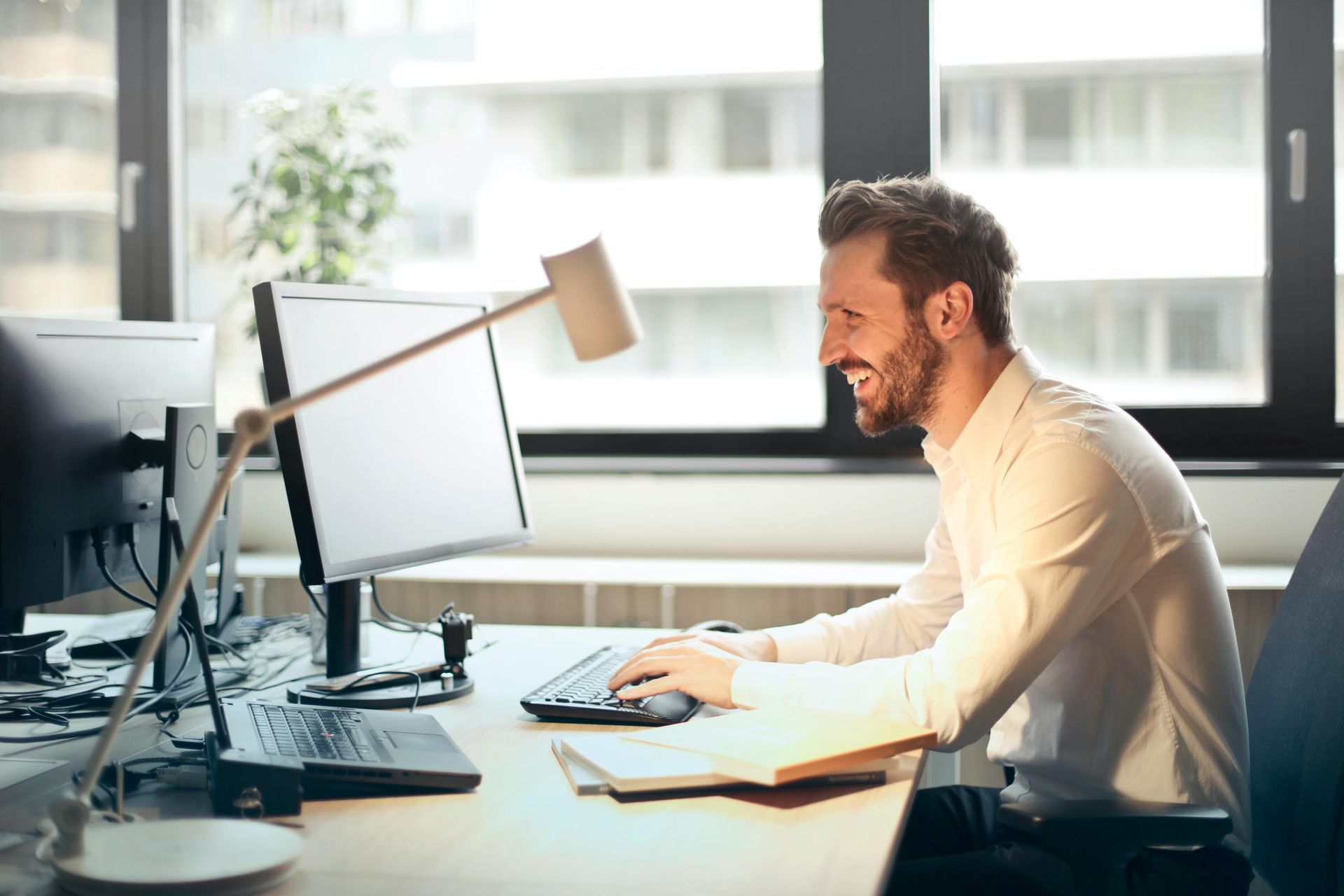 Homem sorrindo, trabalhando no computador em um escritório, com a janela ao fundo