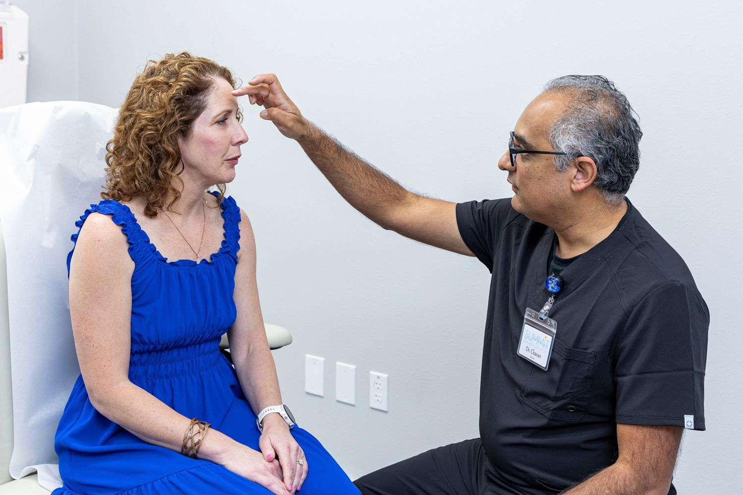 A woman in a blue dress is sitting in a chair talking to a Dr Chavan at Summit Dermatology in Pensacola, Florida