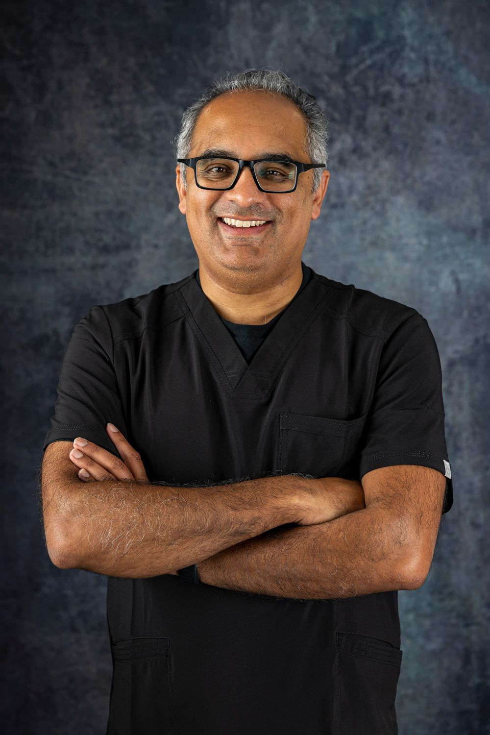 Dr Rahul Chavan in a black scrub top and glasses is standing with his arms crossed and smiling.