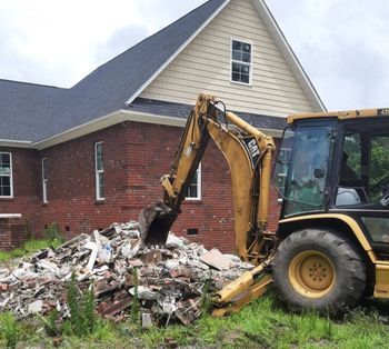 Yellow excavator clearing rubble beside a brick house after demolition