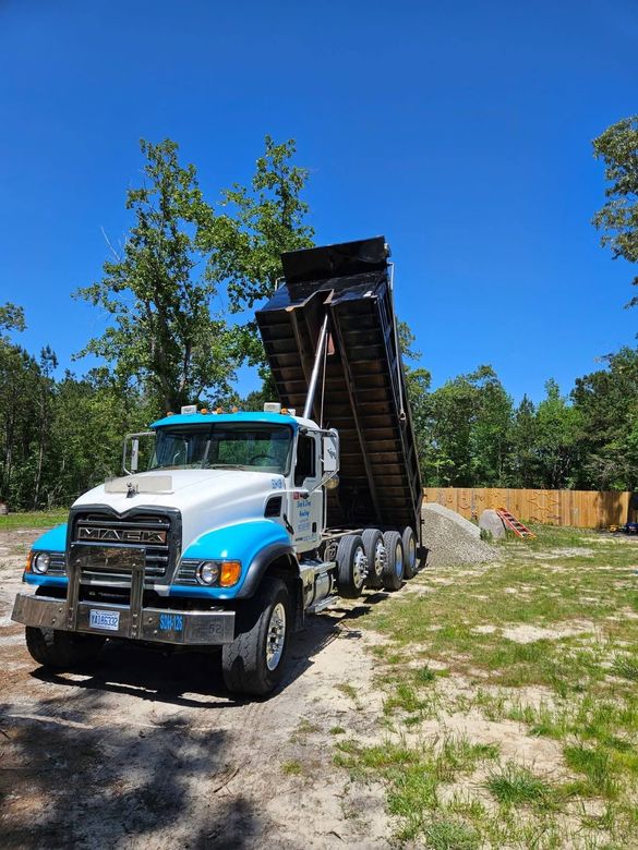 White dump truck lifting a load bed on a sunny gravel lot beside trees and a wooden fence