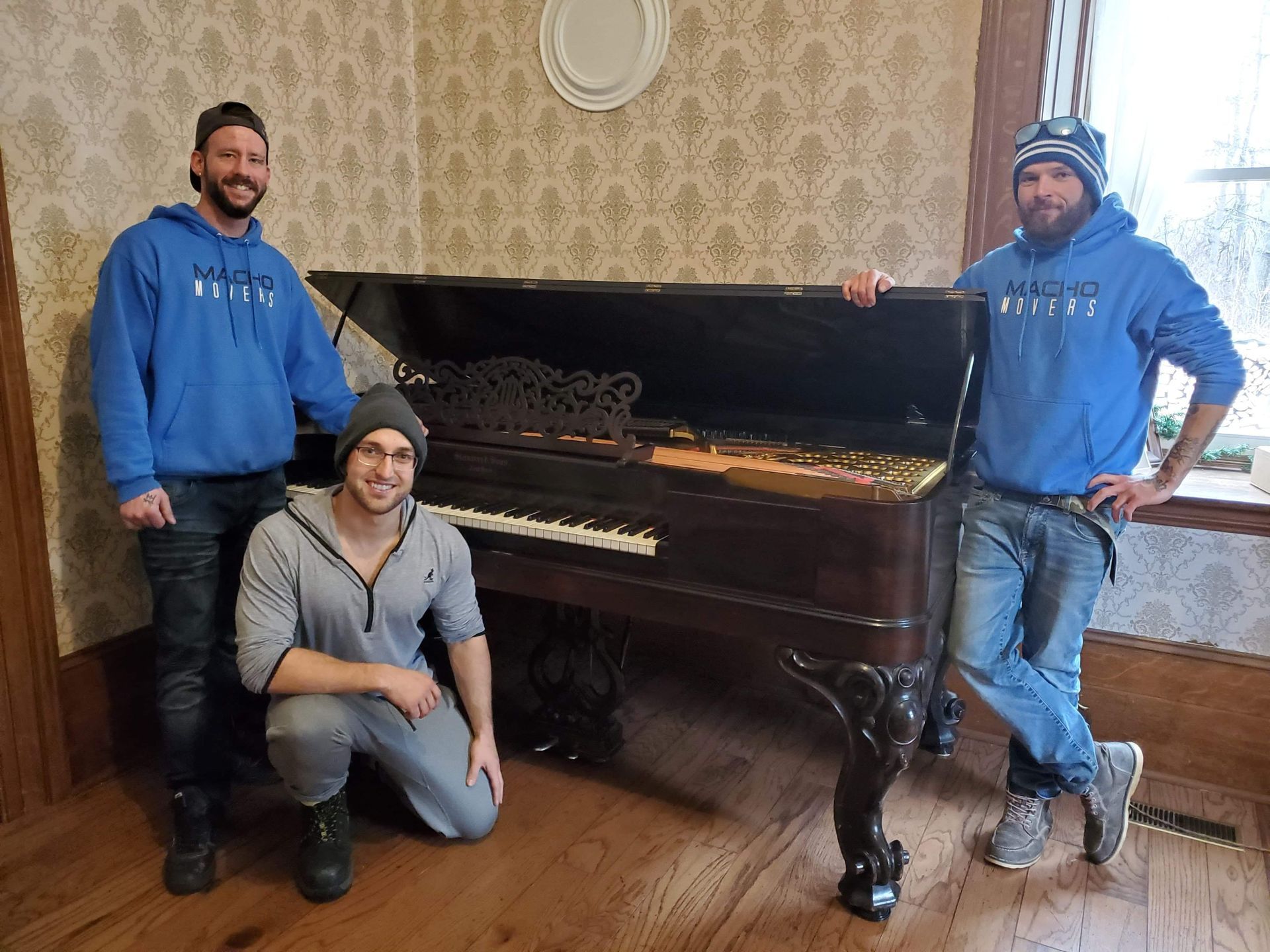 Three people pose with a large, antique grand piano in a room with patterned wallpaper.