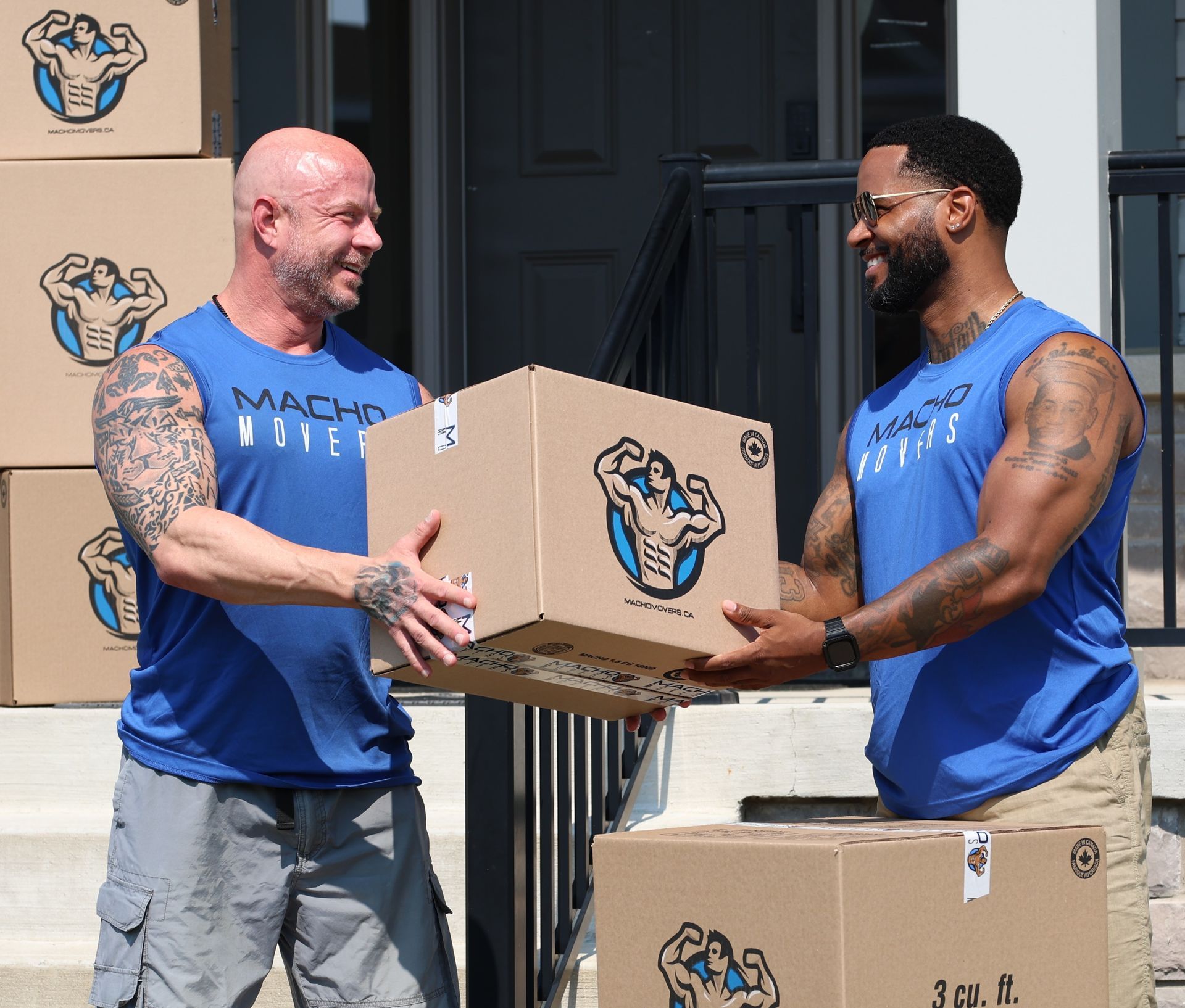 Two men in blue tank tops exchange a cardboard box. Boxes are stacked on steps; one man has tattoos.