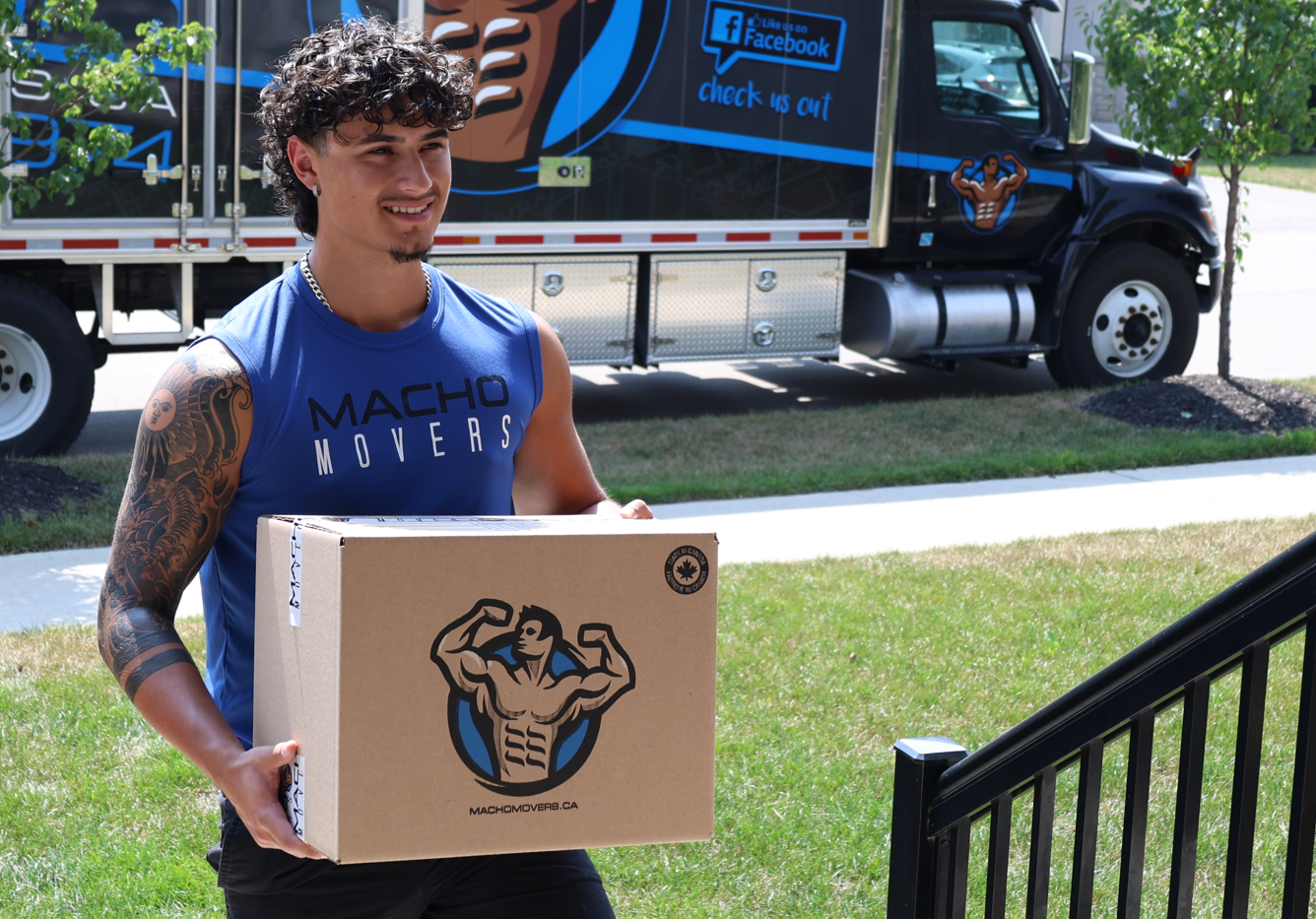 Man in Macho Movers blue shirt, holding box with logo, near delivery truck. Outdoors.