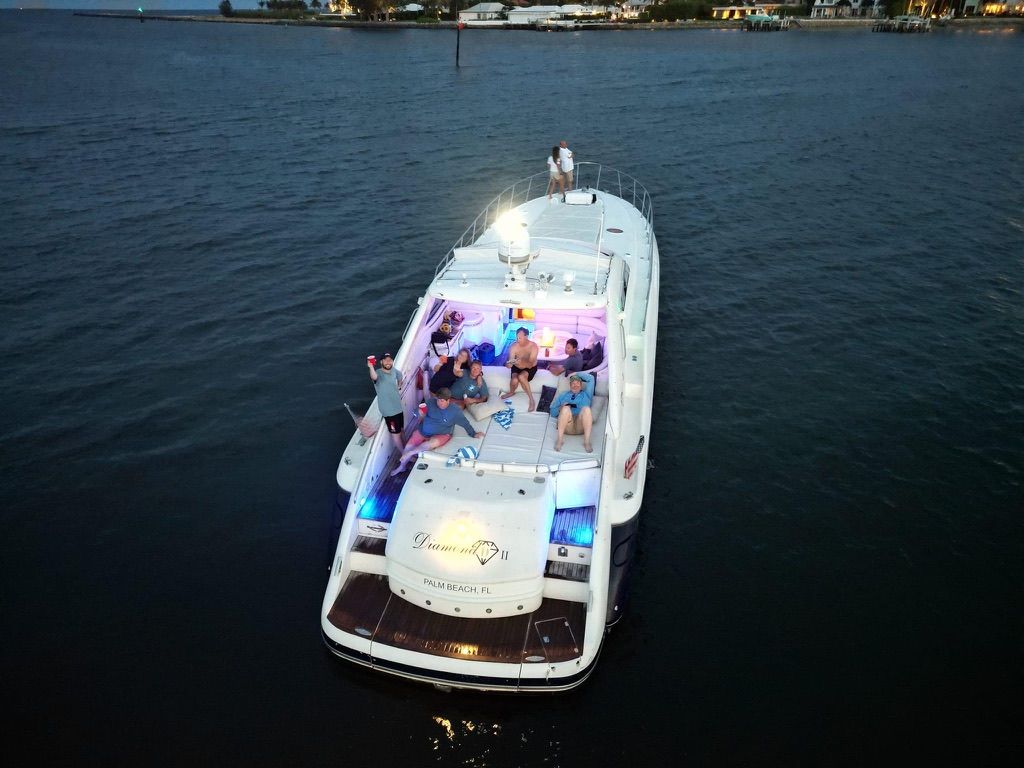 Yacht with people on deck at dusk, lit by colorful lights on calm dark water