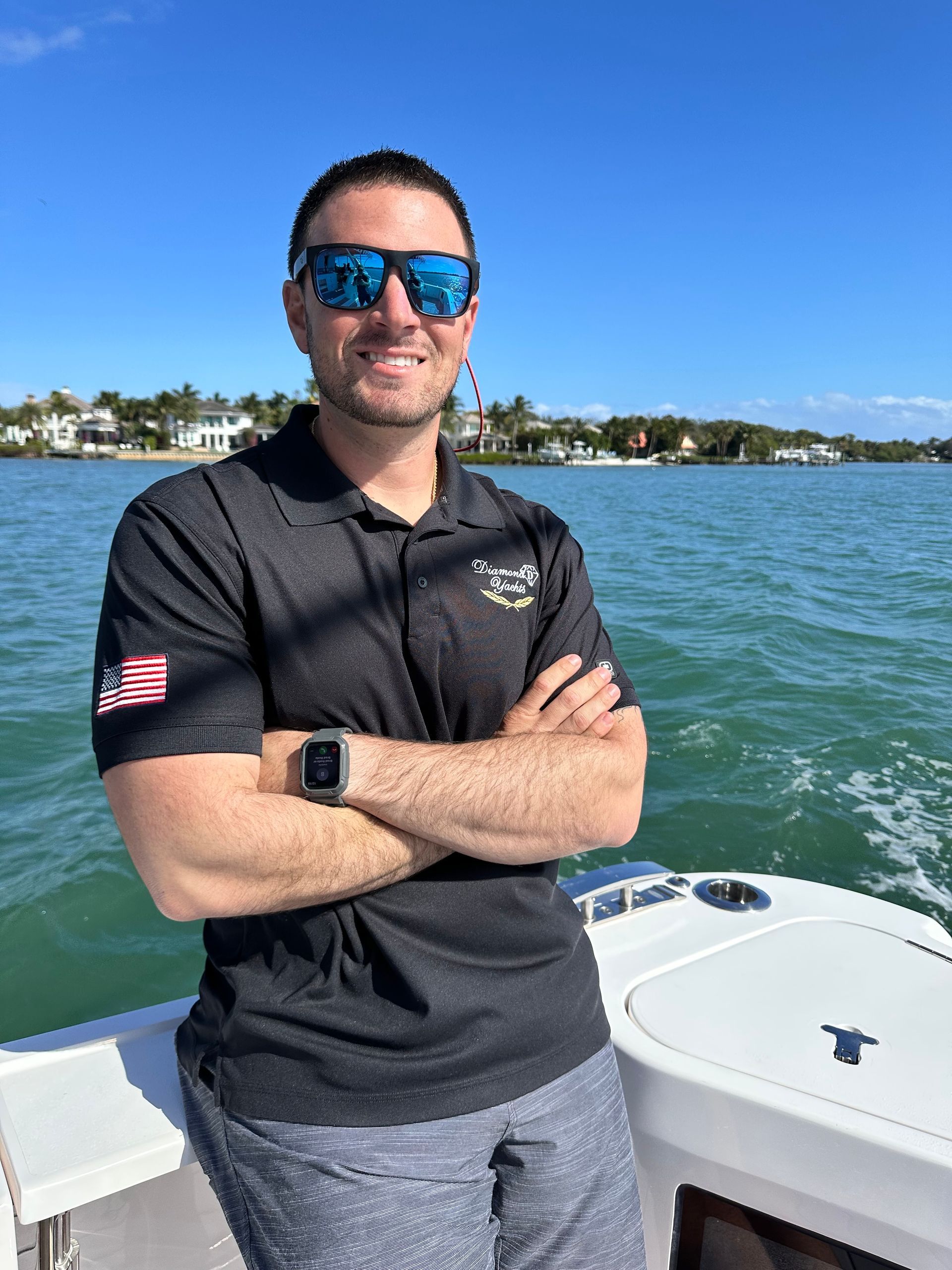 Man on a boat, wearing sunglasses and a black shirt with an American flag patch, arms crossed, water and blue sky background.