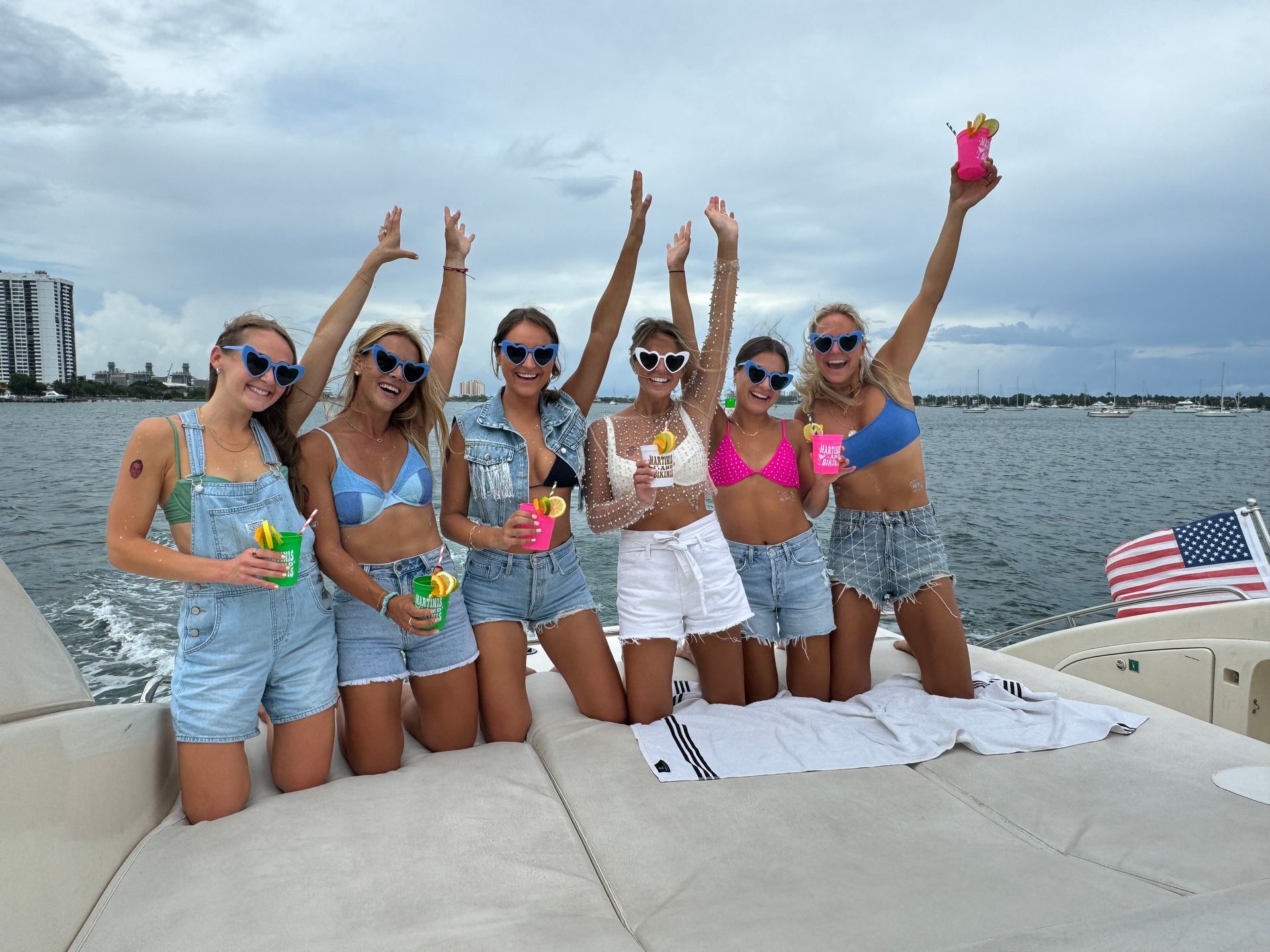 Seven friends in swimsuits posing on a boat deck, holding drinks with a waterfront skyline behind them.