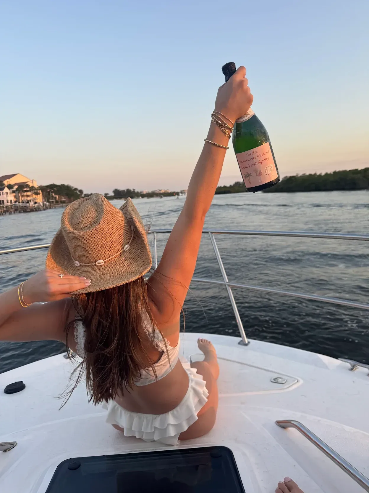 Woman on a boat, holding a champagne bottle up, wearing a straw hat and white bikini.