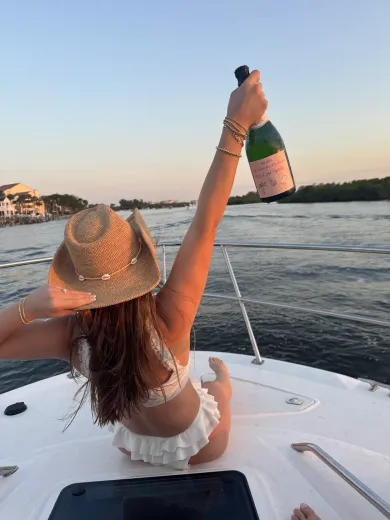 Woman on a boat, holding a champagne bottle up, wearing a straw hat and white bikini.
