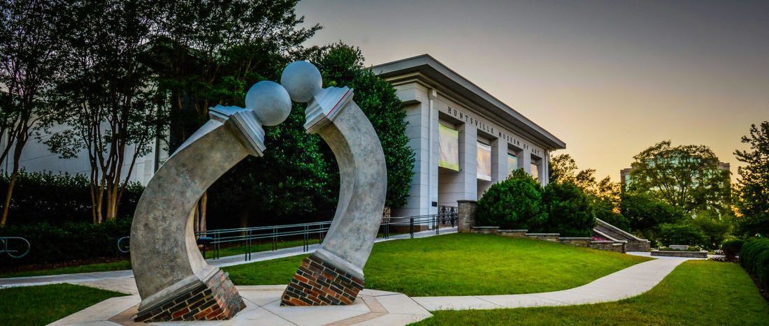 Sculpture in front of a building with a lawn. A winding path leads to a set of stairs. Green grass and sky.