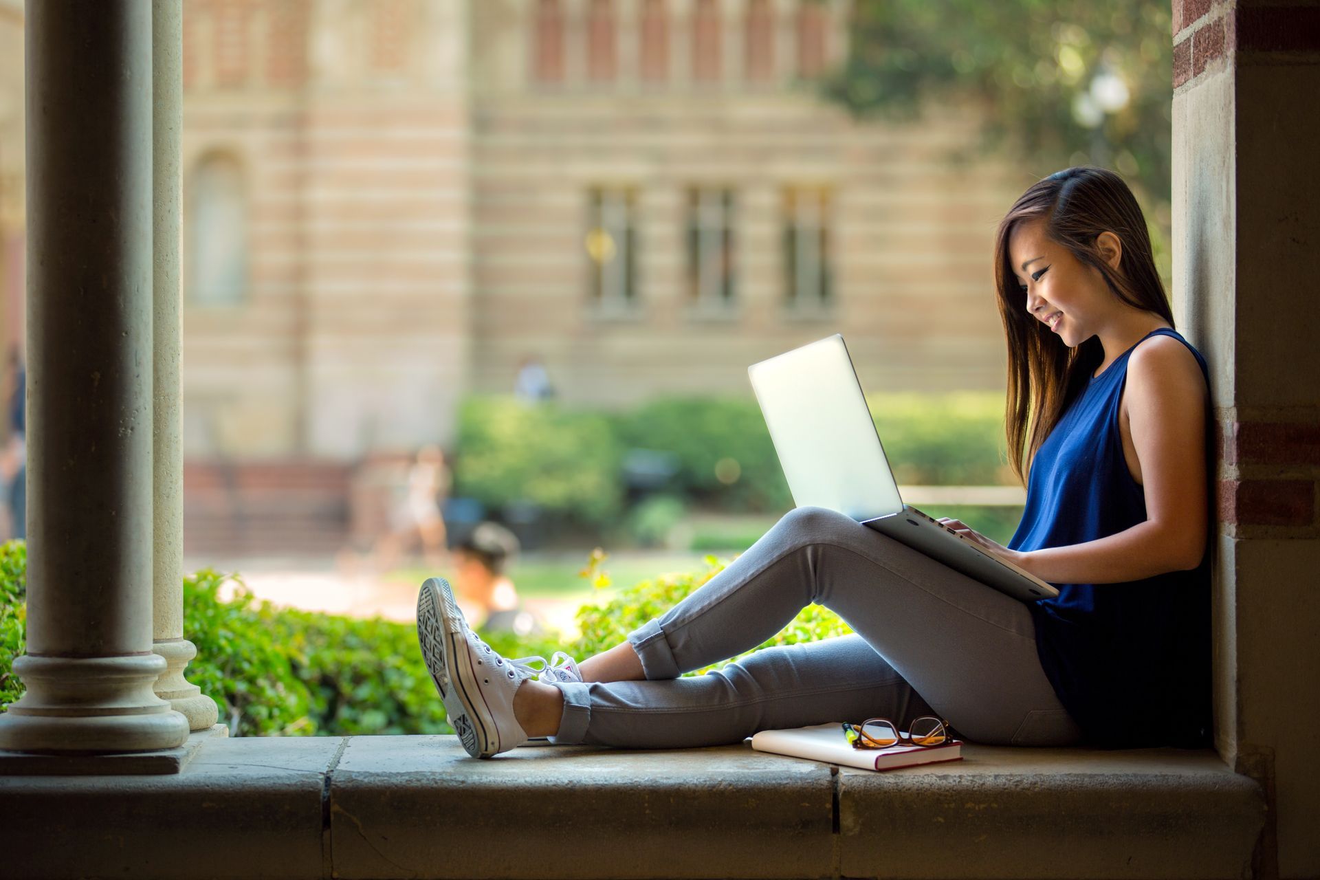 Young woman using a laptop, sitting on a ledge outdoors in front of a building.
