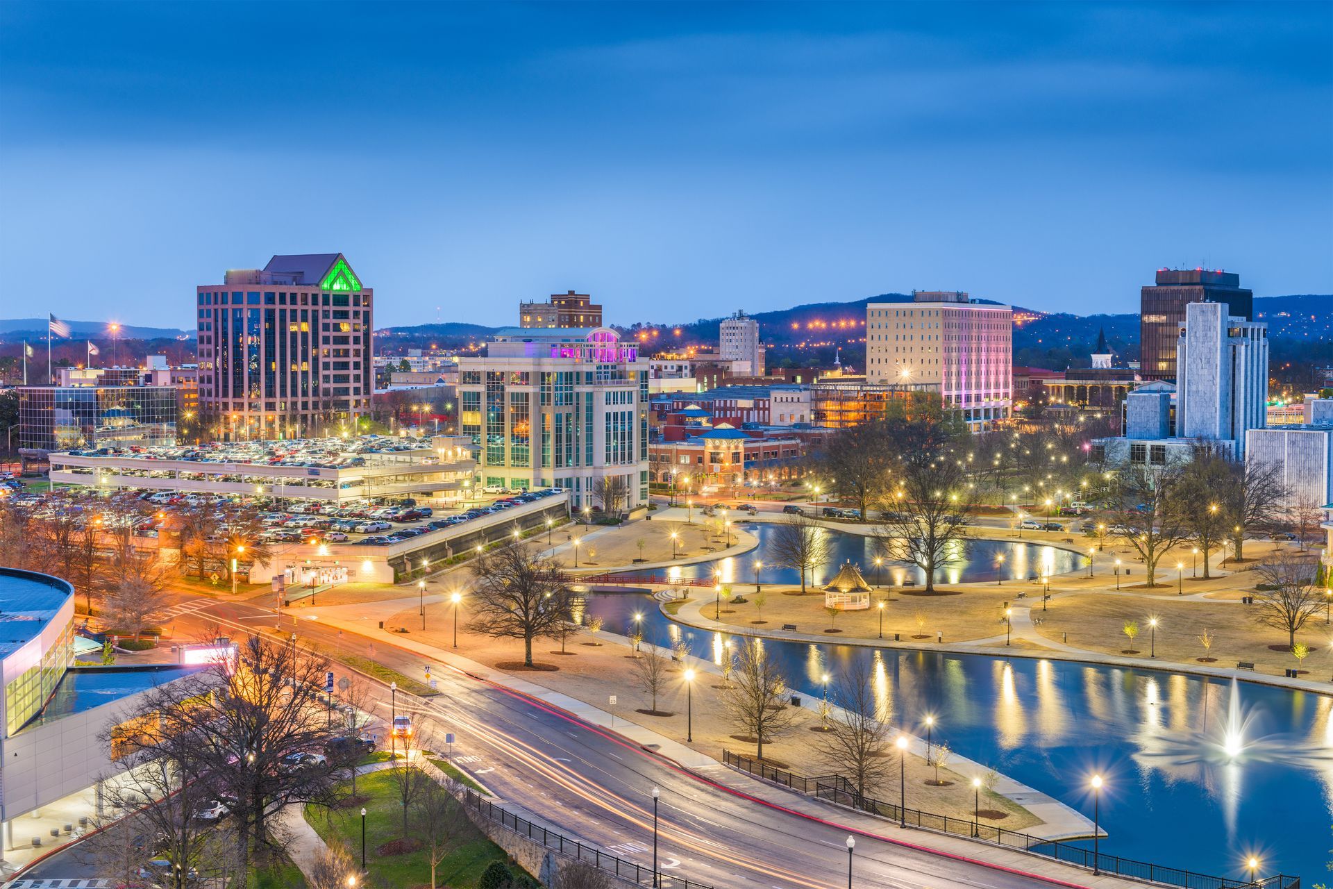 City skyline at dusk with lights reflecting on the water. Buildings of various heights and colors.