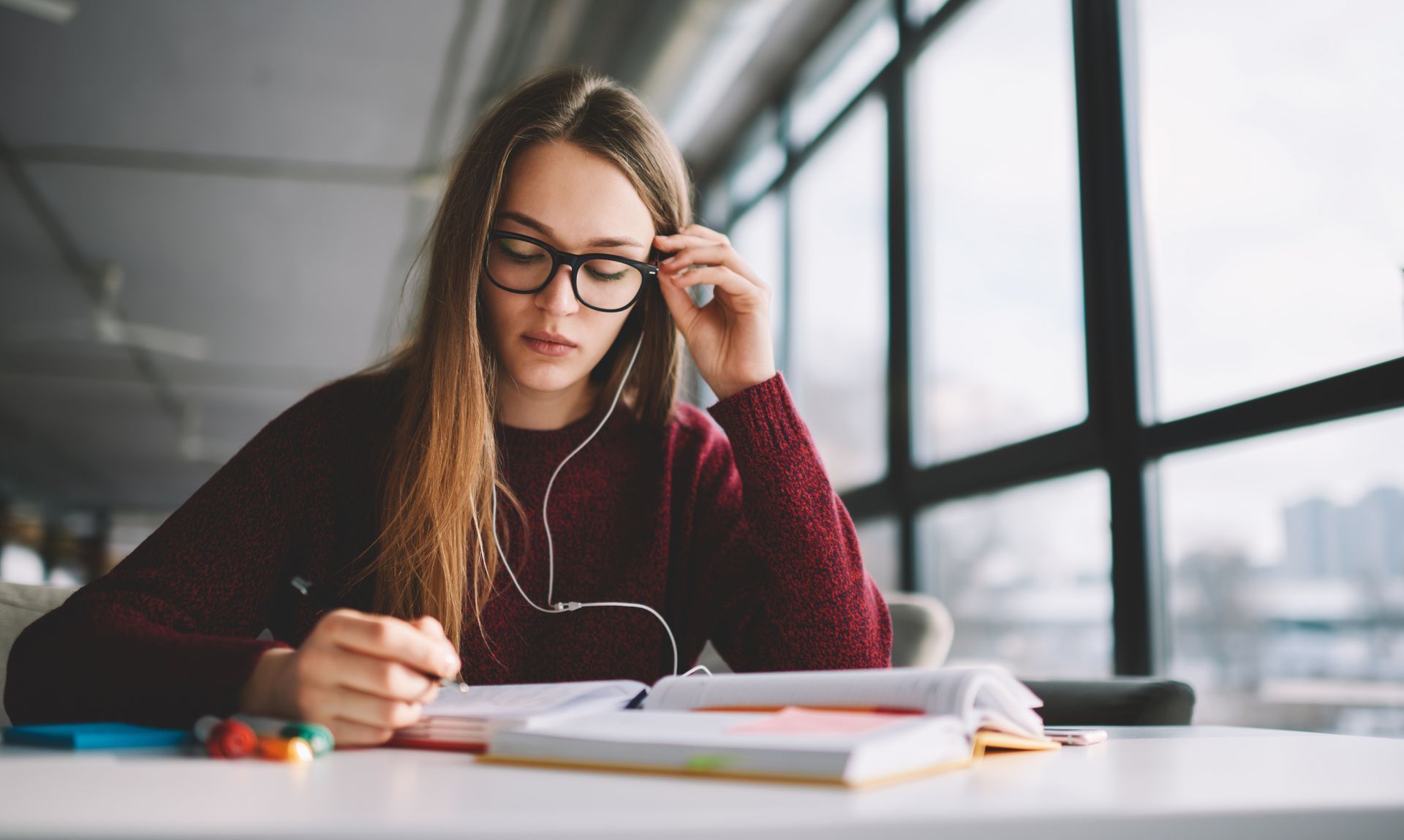 Young person wearing glasses studying at a desk with an open book, window in background.