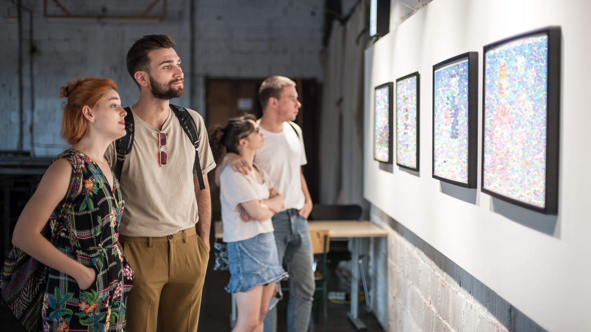 Four people viewing artwork in a gallery. The wall is white with framed abstract art.