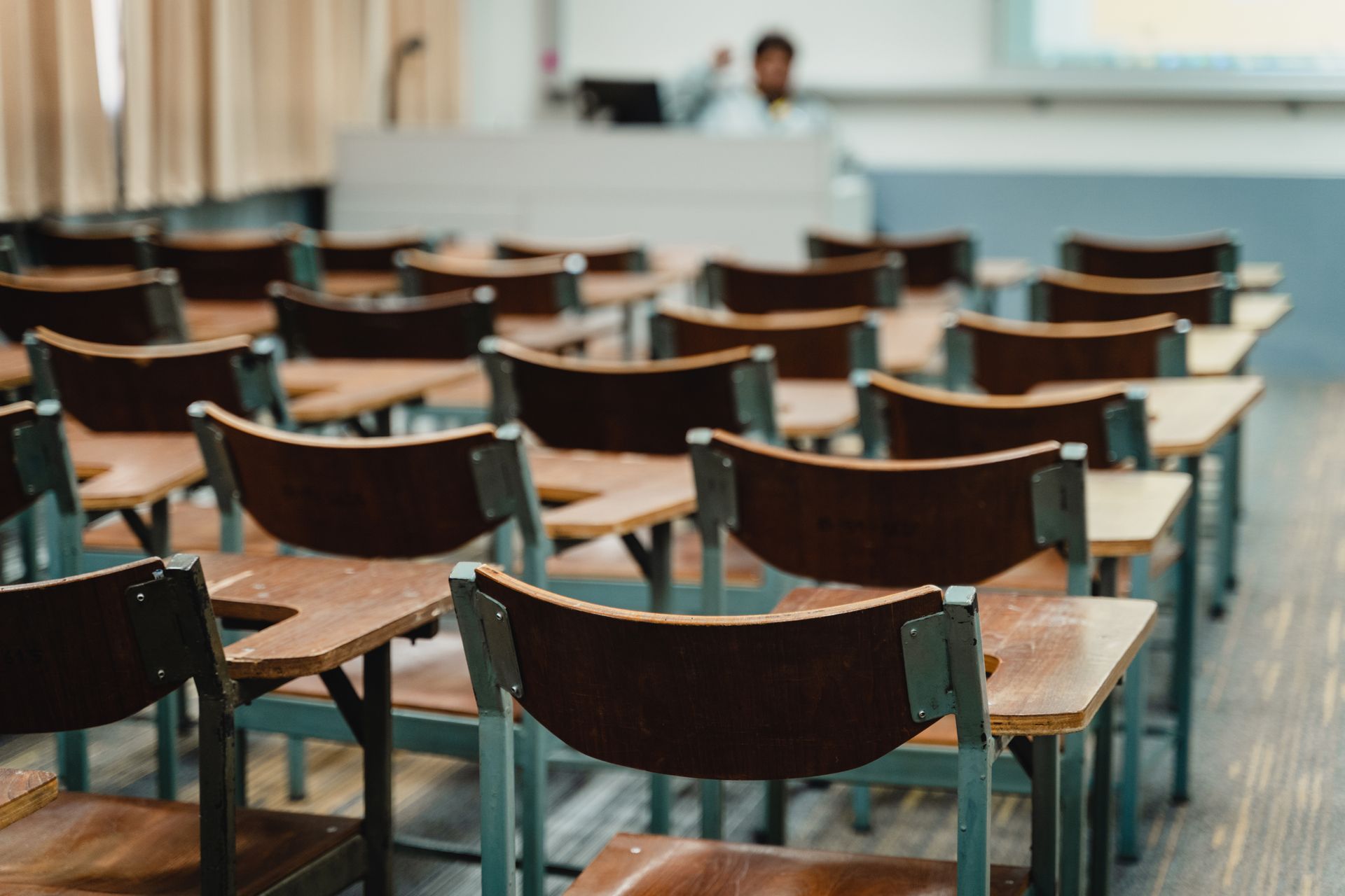 Empty classroom with rows of desks, teacher at front, and tan curtains.