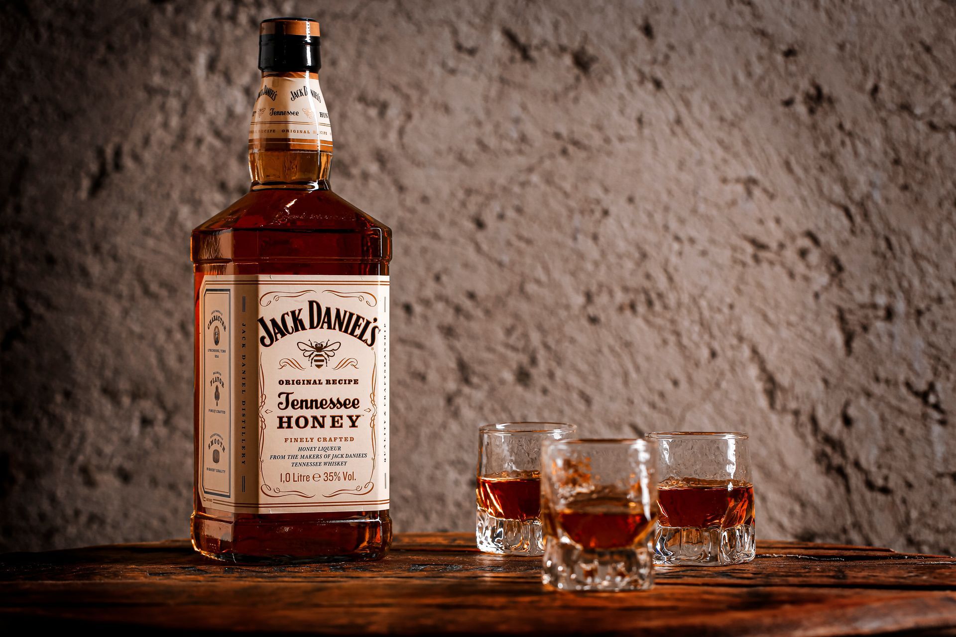 Bottle of Jack Daniel's Honey whiskey with three shot glasses, set against a textured, neutral-toned wall.