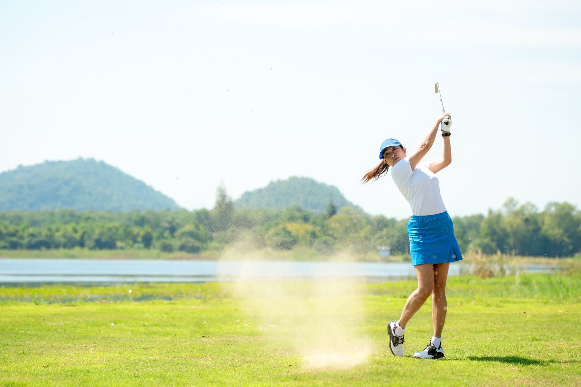Woman swings a golf club on a green course; dust cloud, lake, mountains, blue sky.