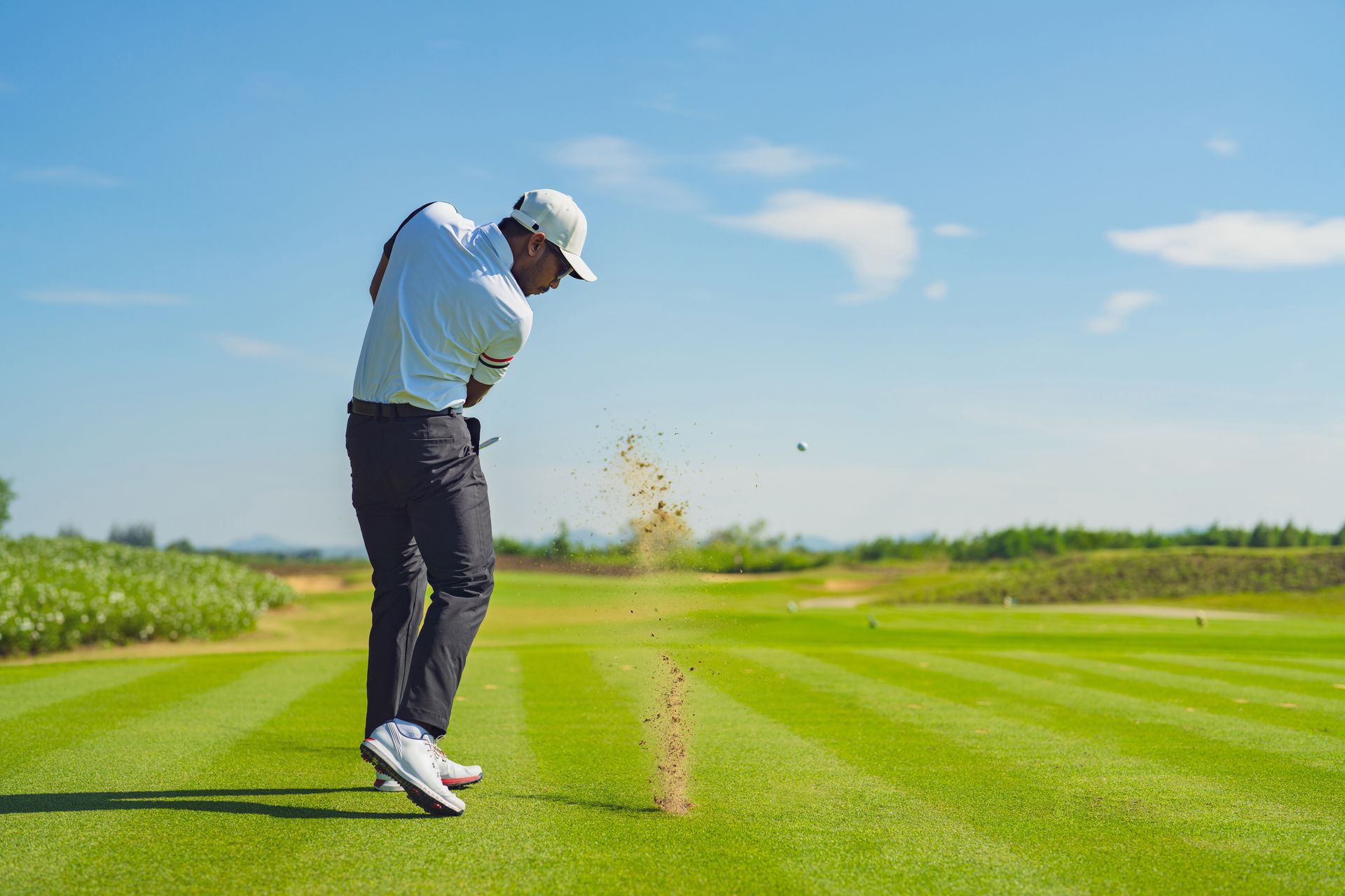 Golfer swinging club on a green course, kicking up sand, sunny day.