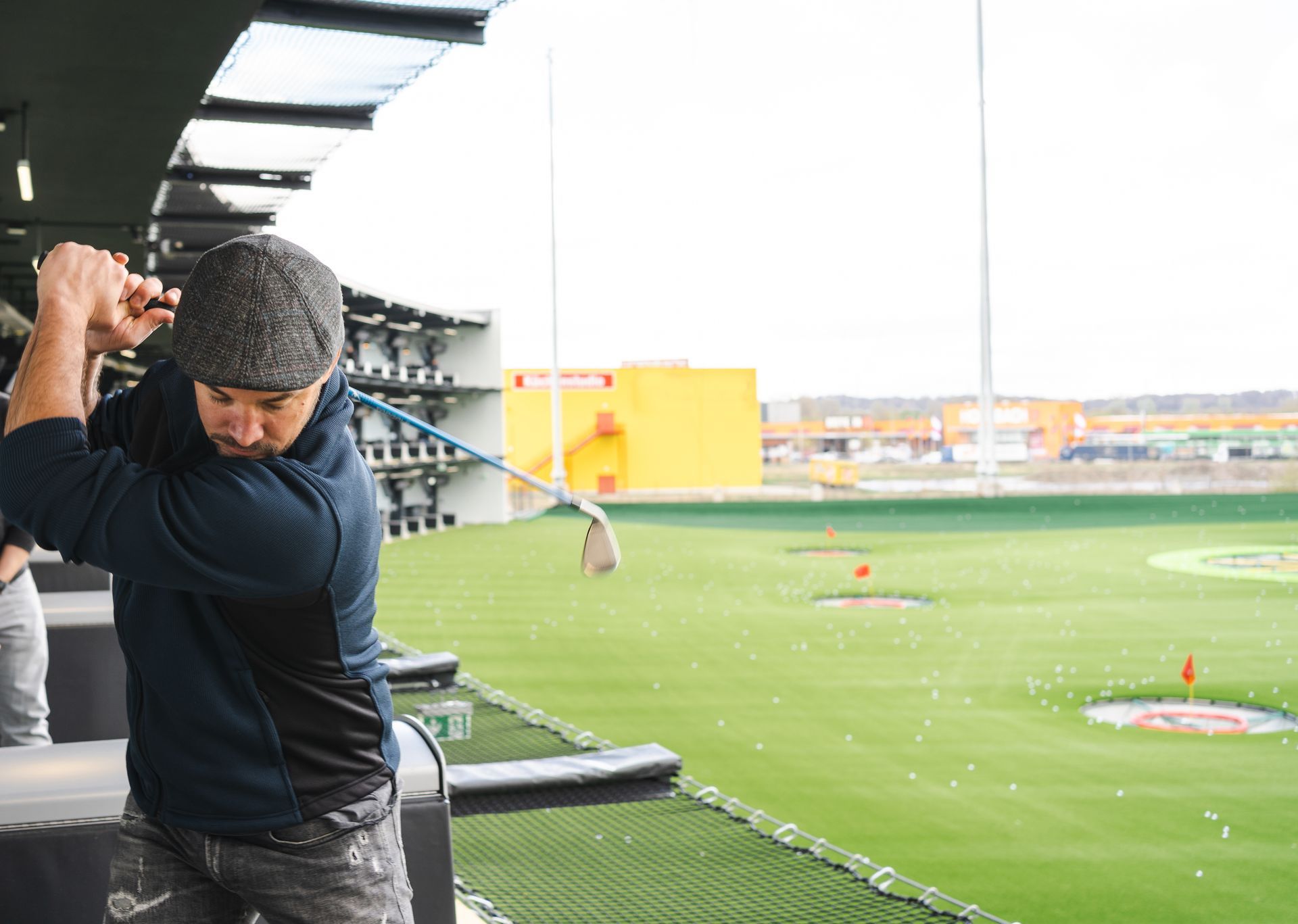 Man swinging a golf club at a driving range, with green targets and a yellow building in the background.