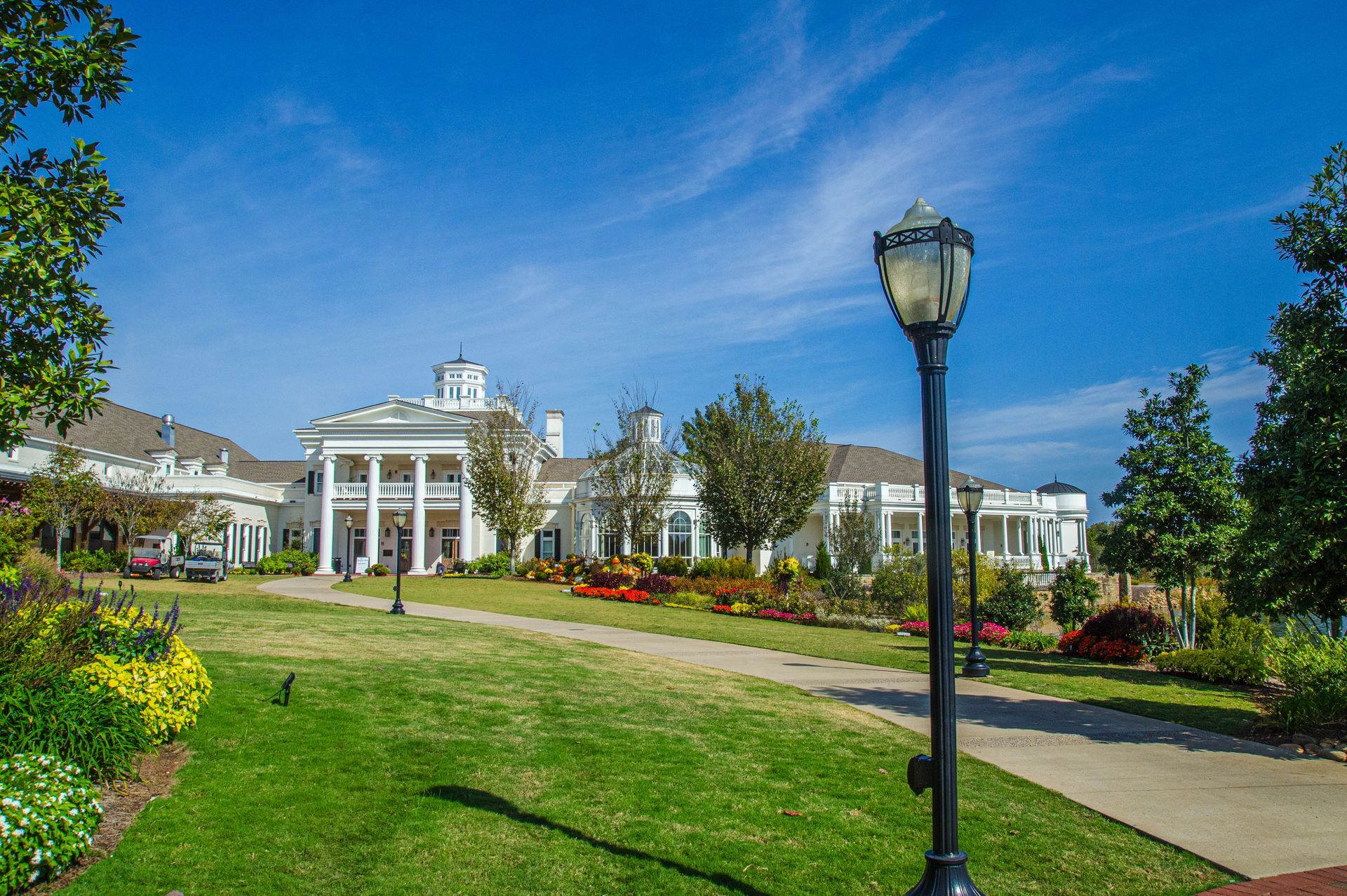 White building with columns on a bright day. Green lawn, flower beds, lamp post, blue sky.